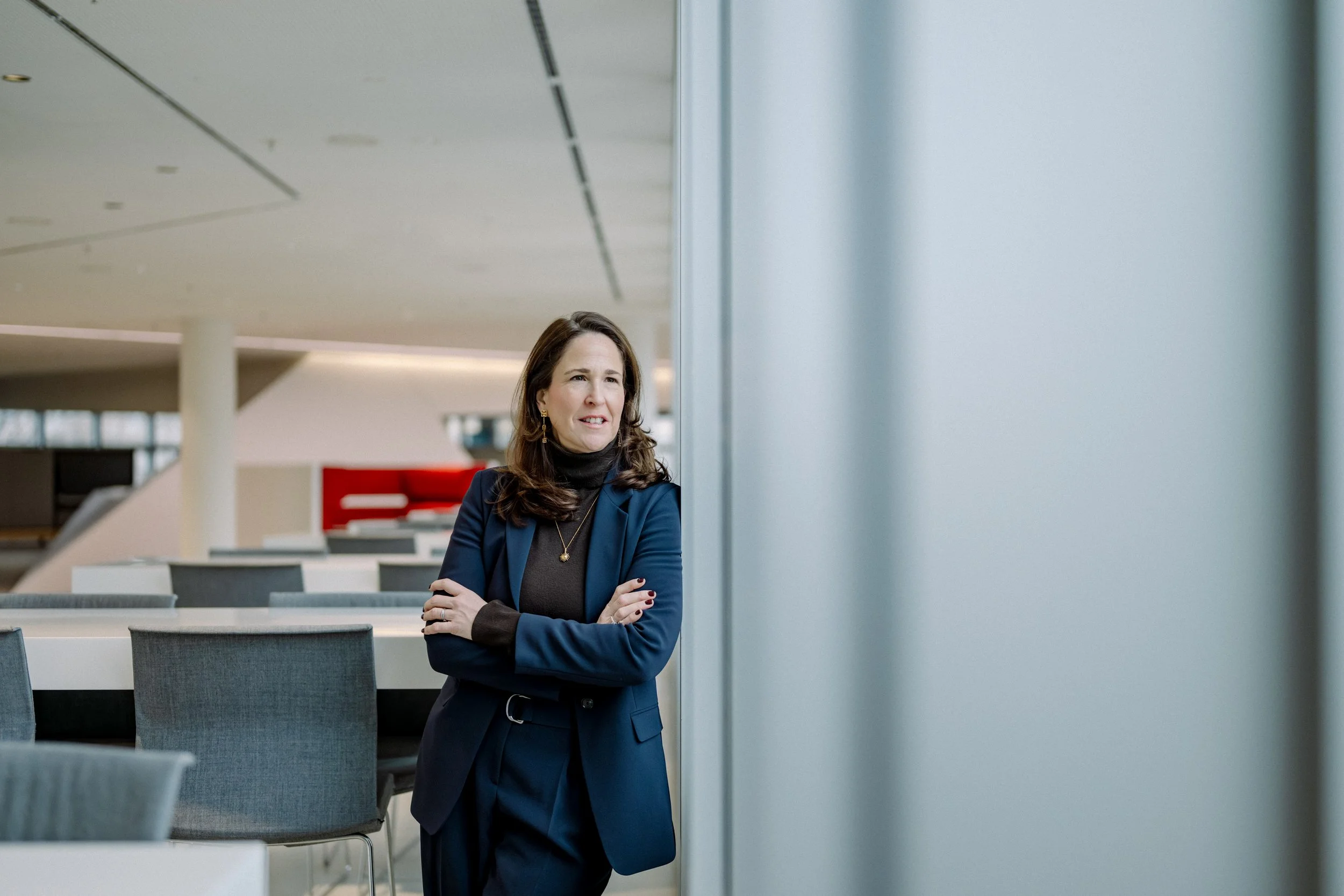 Frau in dunklem Blazer steht in einem modernen Wartebereich oder einer Lobby, blickt nach oben und lächelt leicht, umgeben von leeren Tischen und Stühlen.