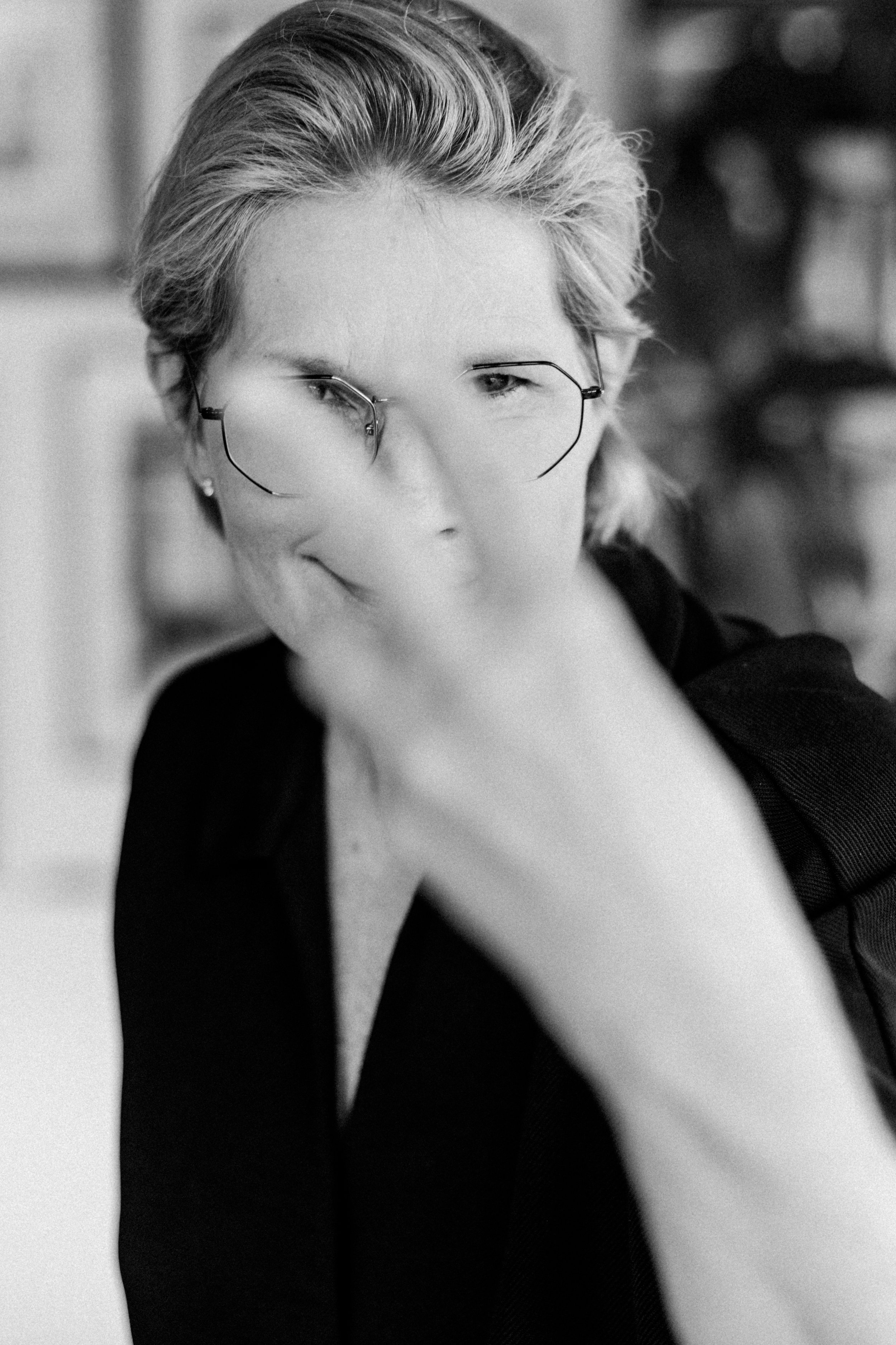 Schwarz-weiß-Foto einer Frau mit kurzen, gestylten Haaren, die in eine Richtung schaut, trägt randlose Brille und eine dunkle, ärmellose Kleid. Der Hintergrund ist unscharf.