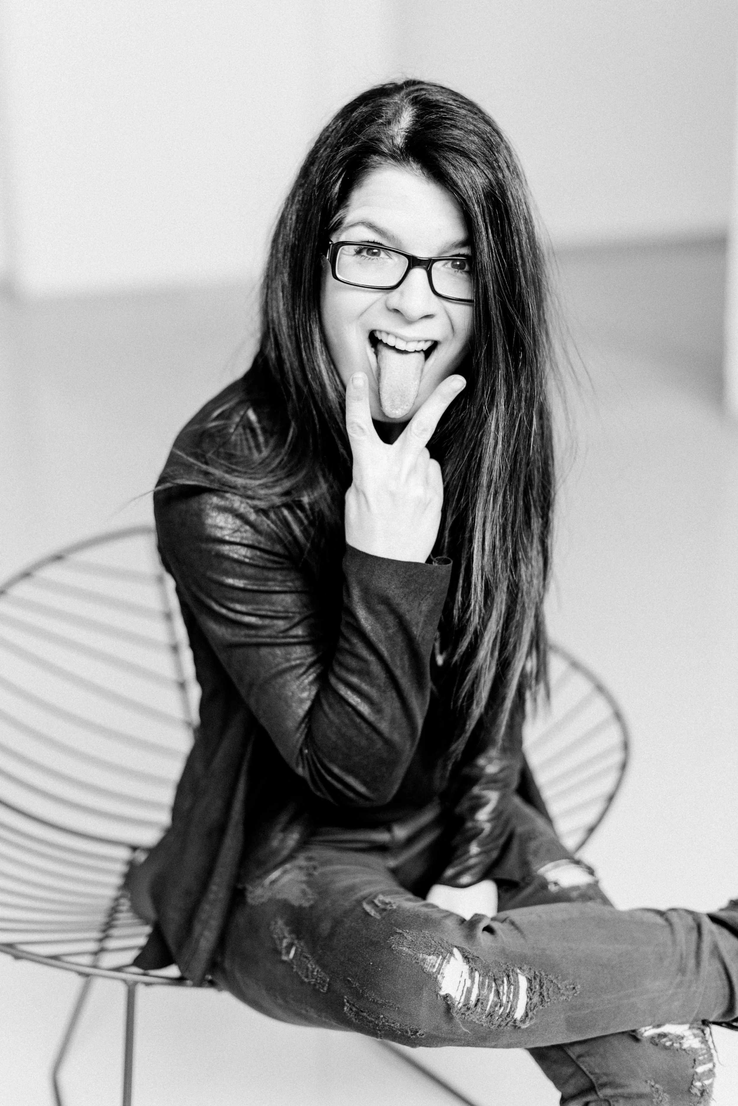 Schwarz-Weiß-Foto einer lächelnden jungen Frau mit Brille, die die Zunge rausstreckt und eine peace-Zeichen-Geste macht, während sie auf einem modernen Stuhl sitzt.