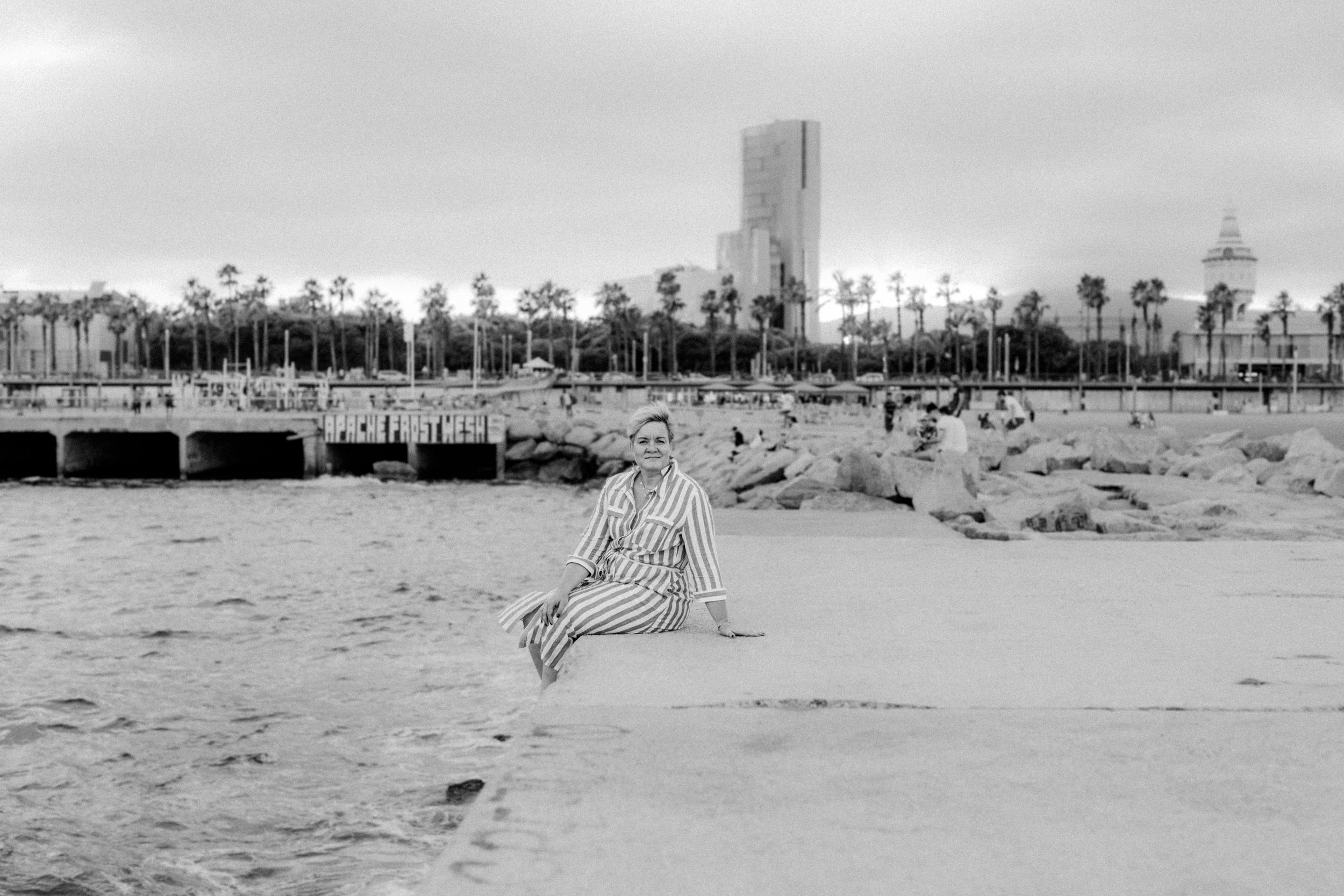 Eine Frau in einem gestreiften Kleid sitzt auf einem Felsen am Strand, im Hintergrund befinden sich Palmen, Gebäude und eine Stadtansicht.