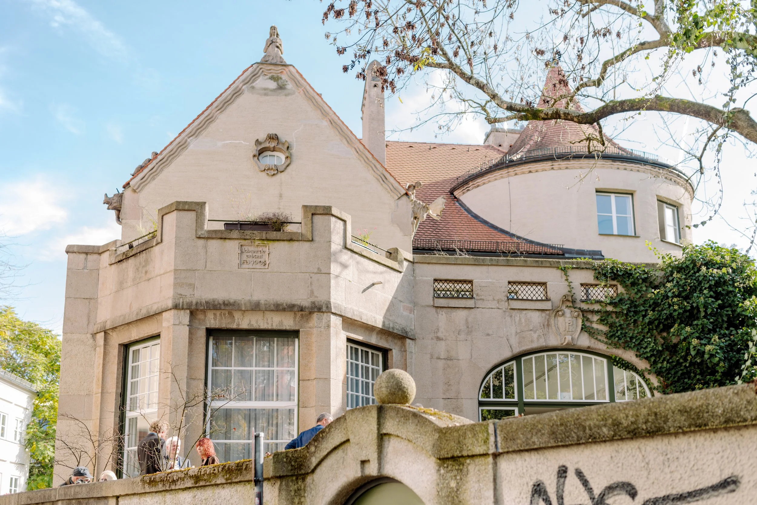 Ein Gebäude im Jugendstil mit runden Erkern, Dachgauben und kunstvollen Verzierungen, umgeben von Bäumen, im Vordergrund eine Mauer mit Figuren und Menschen. Sandsteinstrukturen, große Fenster und ein Mauerstück mit Schriftstück.