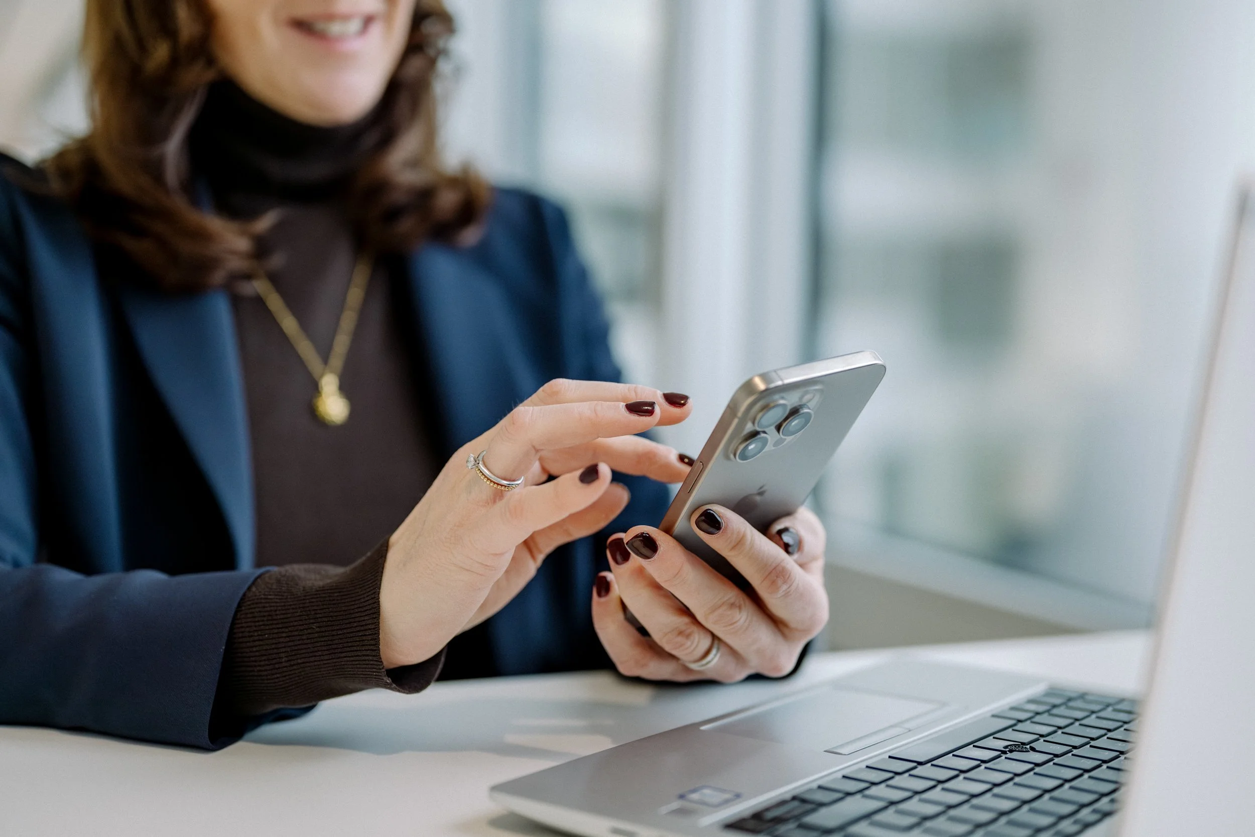 Eine Frau benutzt ihr Smartphone an einem Schreibtisch, ein Laptop ist geöffnet vor ihr. Sie trägt dunkle Kleidung und eine goldene Halskette.