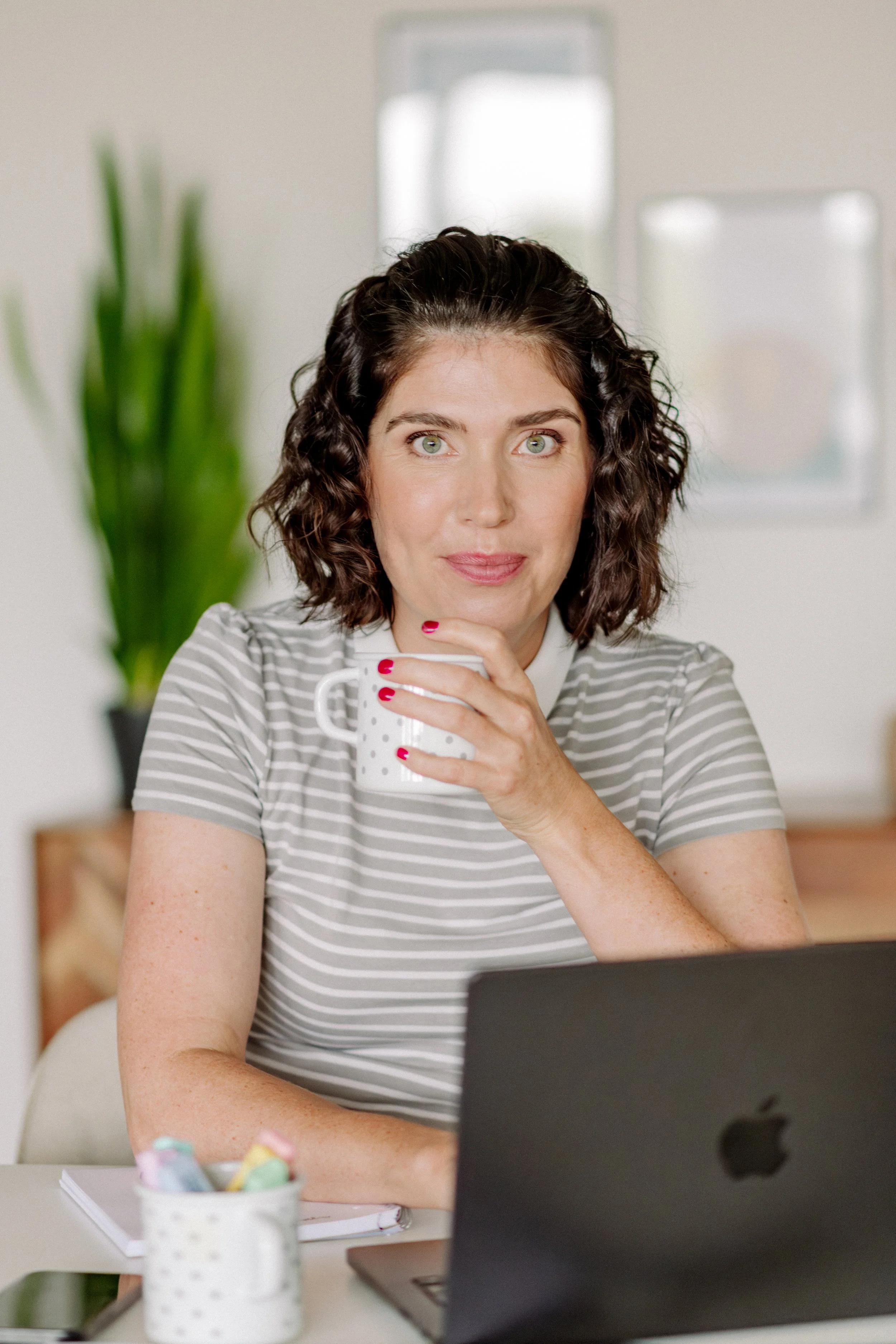 Eine Frau mit braunen, lockigen Haaren und grünen Augen sitzt vor einem Laptop. Sie trägt ein gestreiftes T-Shirt und hält eine Tasse in der Hand. Im Hintergrund sind ein Fenster und eine Pflanze sichtbar.