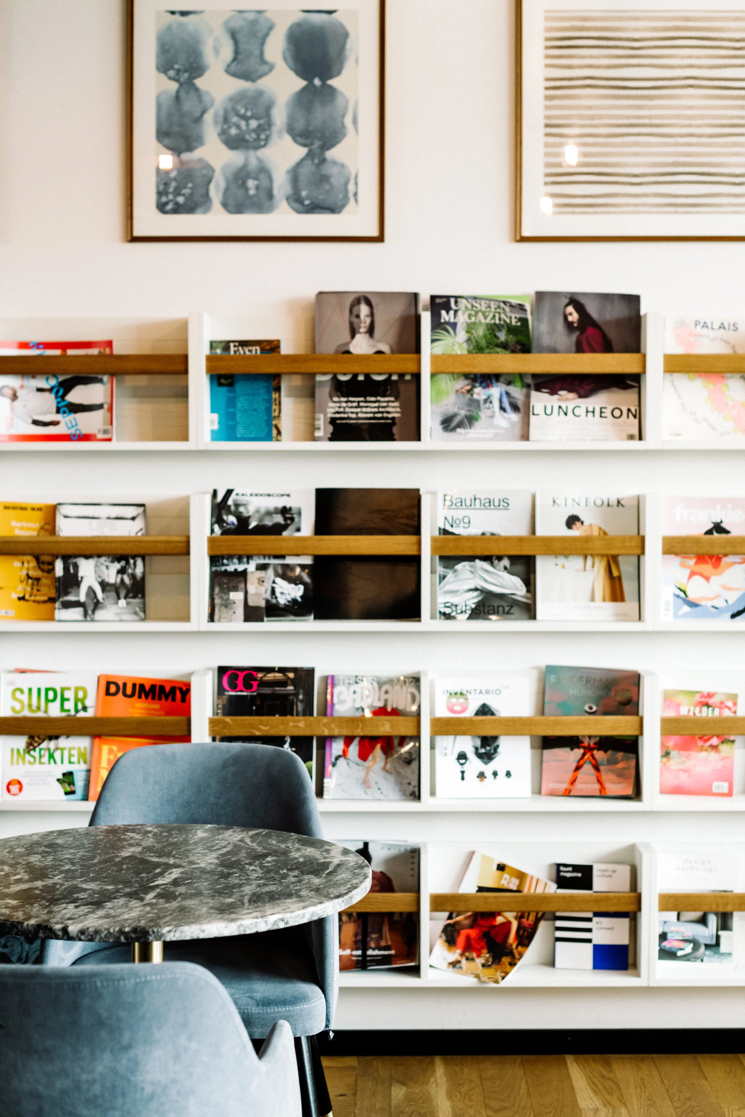 Buchregale mit Zeitschriften und Büchern an einer weißen Wand mit zwei Kunstwerken darüber, vor einem runden Steintisch und grauen Stühlen in einem Raum.