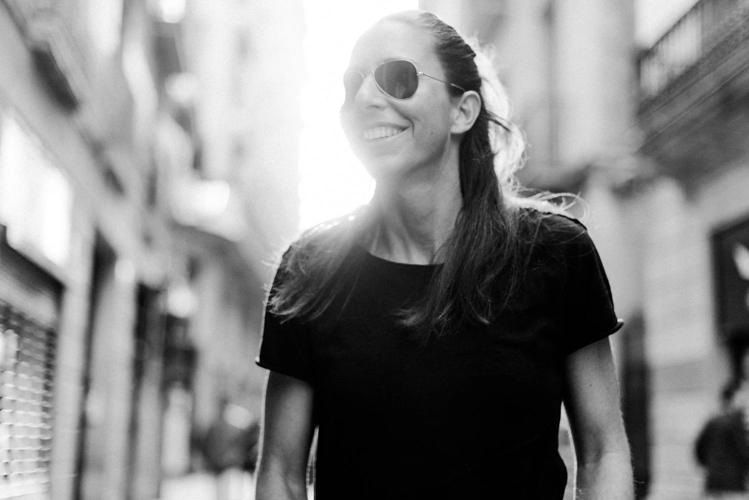 Eine lachende Frau mit dunklem Haar, Sonnenbrille und schwarzem T-Shirt geht in einer Stadtstraße, im Hintergrund Gebäude, bei Sonnenlicht. Schwarz-Weiß-Fotografie.