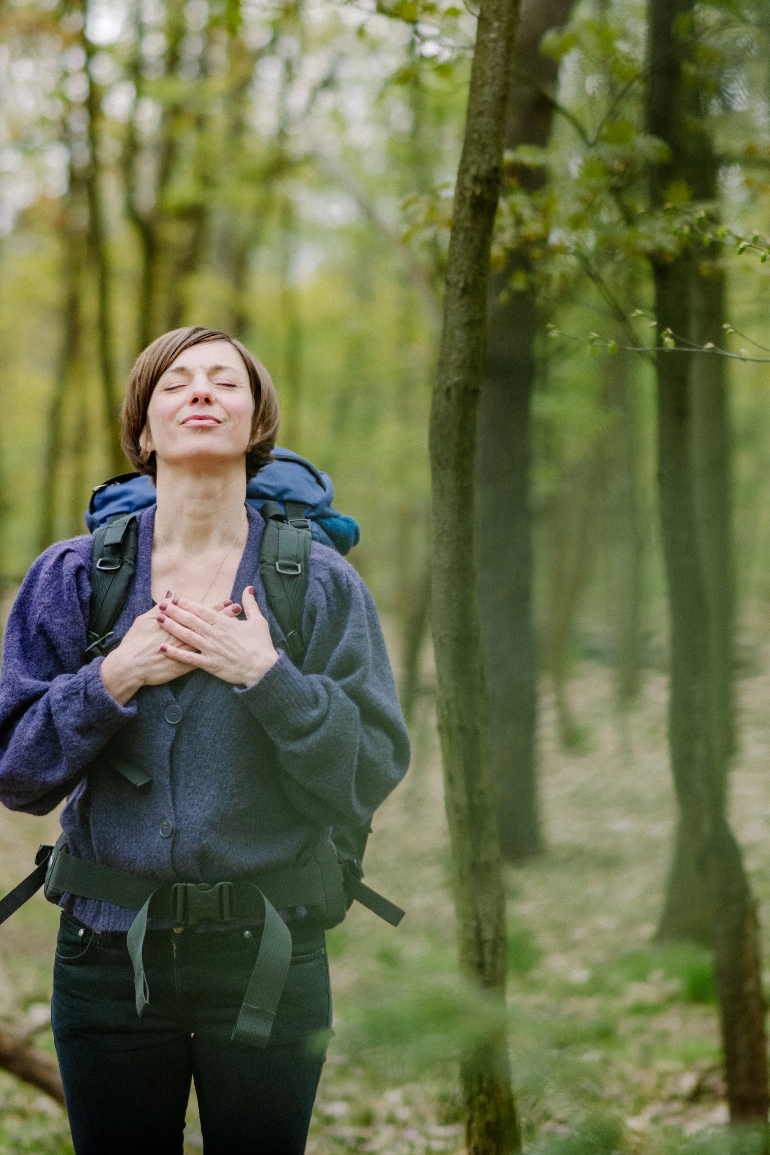 Eine Frau mit kurzen braunen Haaren trägt einen blauen Rucksack und eine graue Jacke, steht in einem grünen Wald und zeigt Andacht oder Freude mit zusammengelegten Händen auf die Brust, die Augen geschlossen.