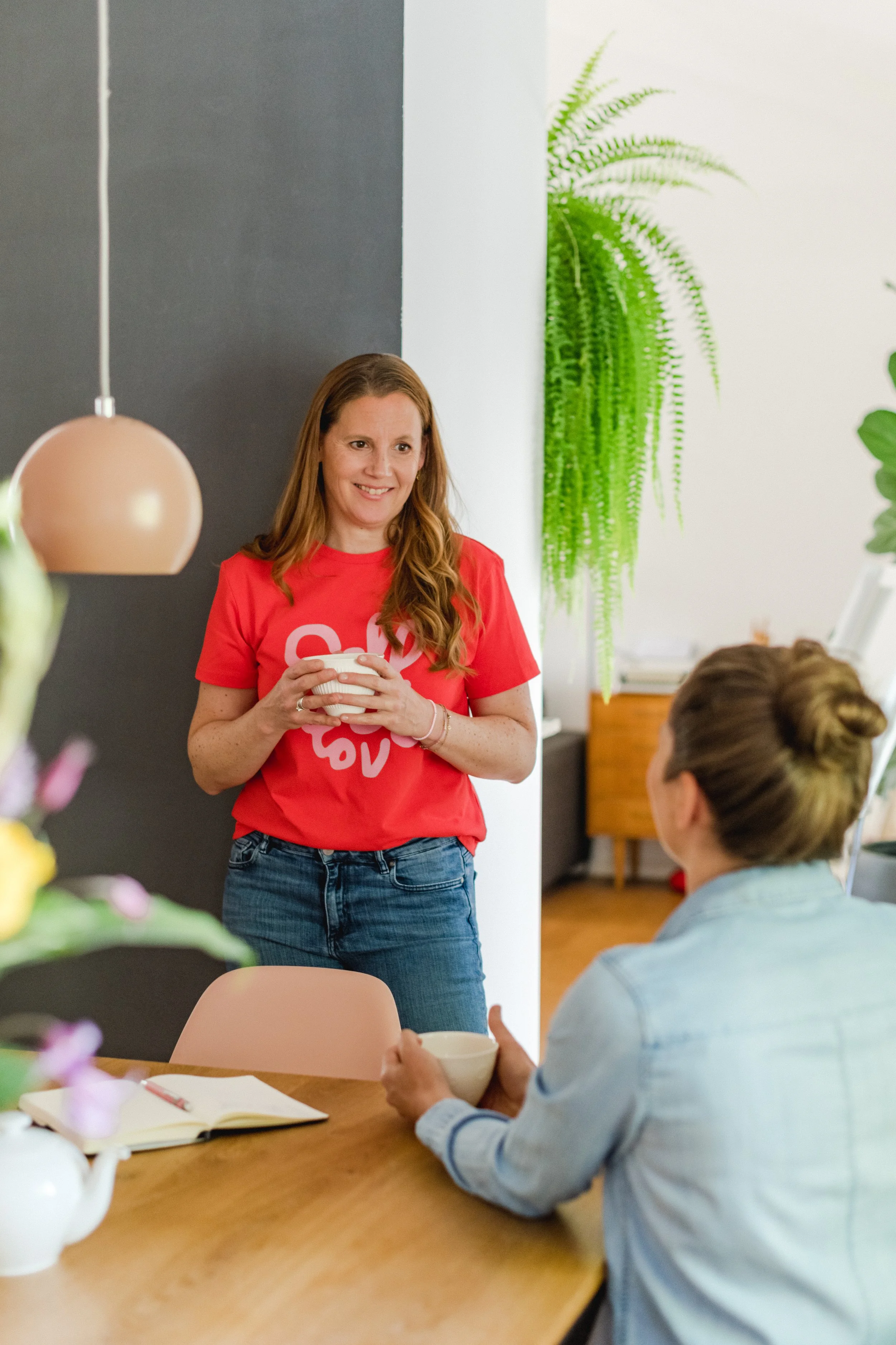 Eine Frau mit langen, braunen Haaren, die ein rotes T-Shirt trägt, steht in einem Raum, der an eine Küche erinnert. Sie hält eine Tasse in den Händen und lächelt. Zwei weitere Personen sitzen an einem Holztisch, einer trägt eine hellblaue Jeansjacke 
