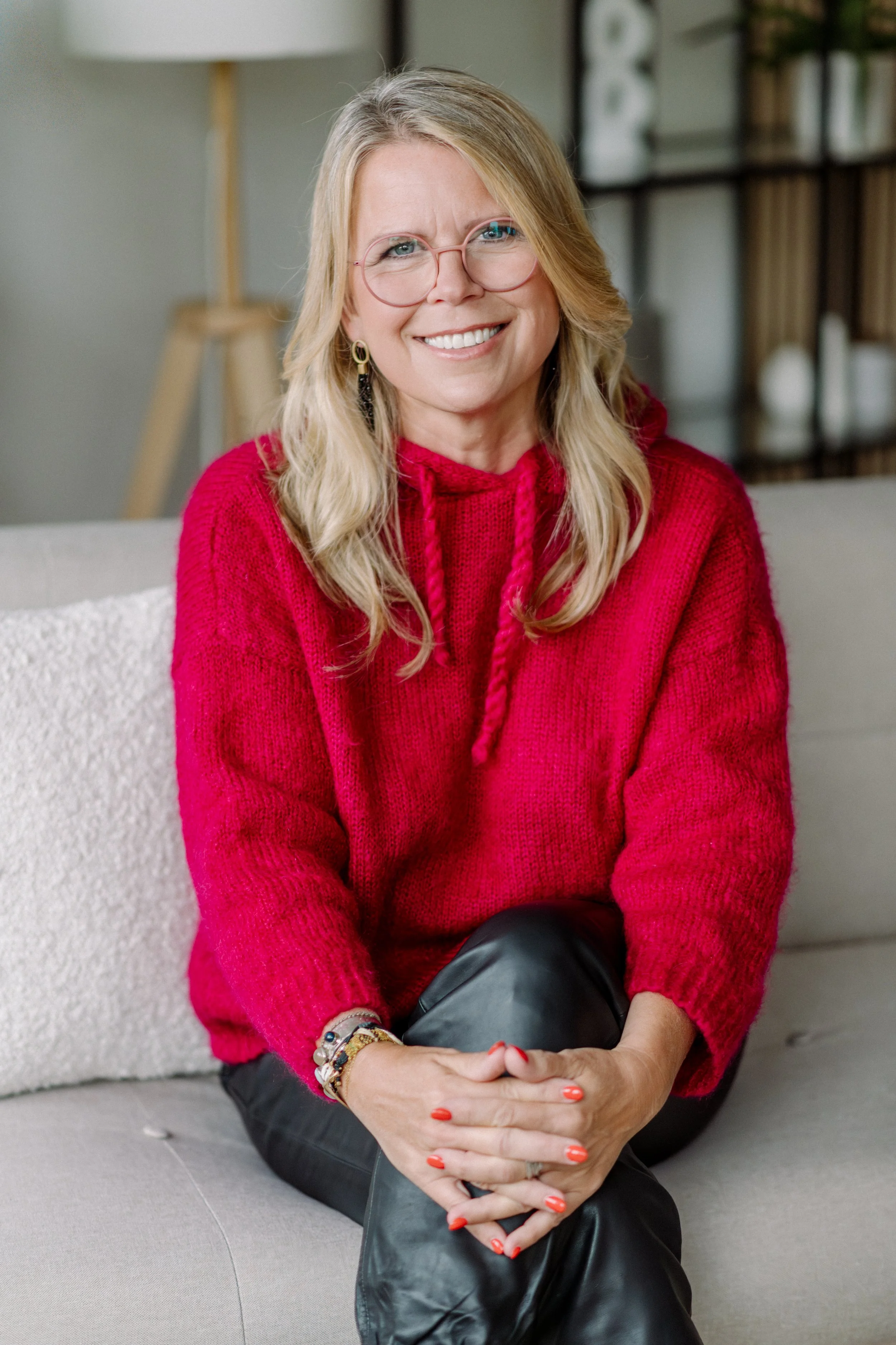 Eine Frau mit blonden Haaren, Brille und rotem Pullover sitzt auf einer Couch und lächelt. Sie trägt mehrere Armbänder und hat rote Nagellacke.