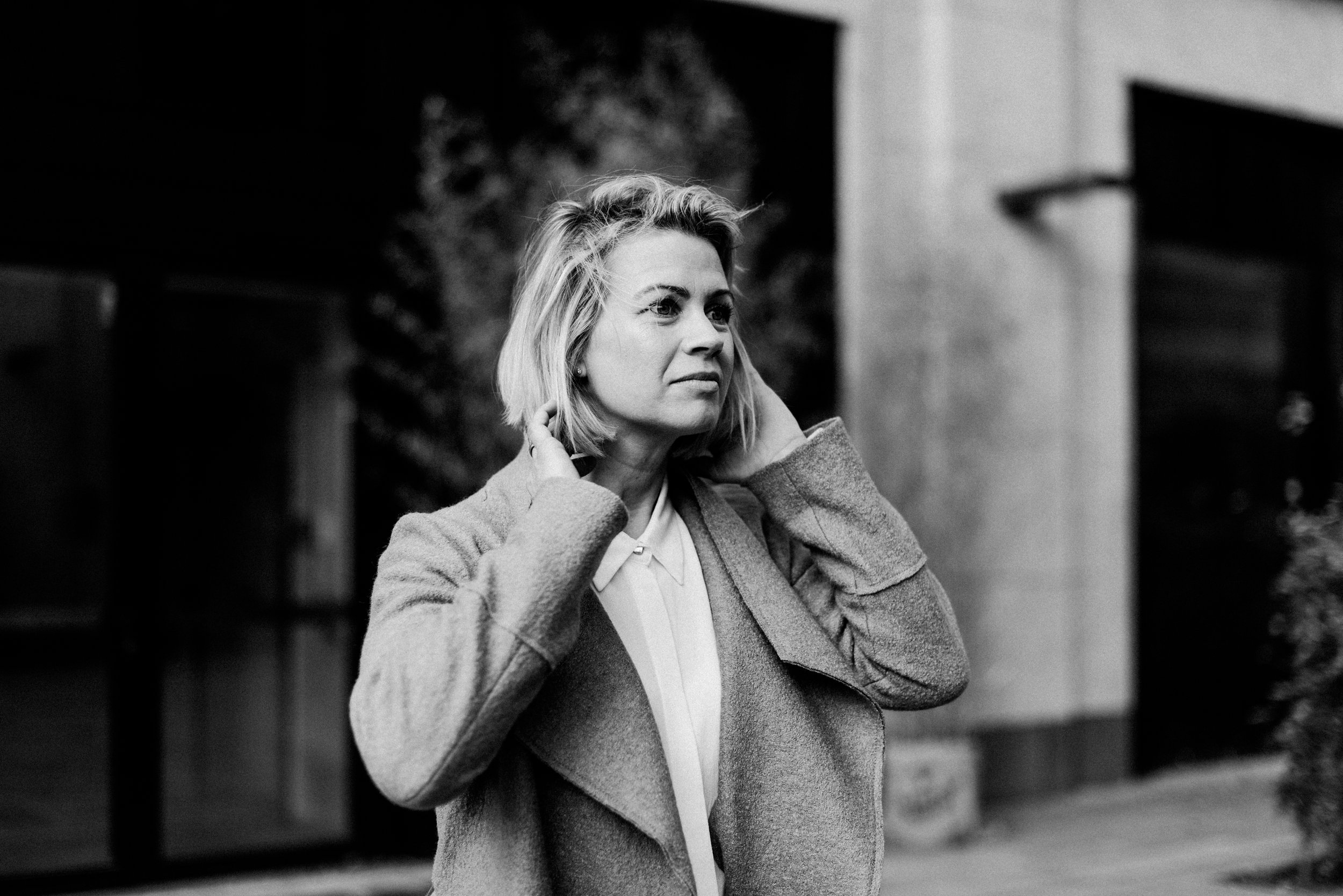 Schwarz-weiß-Foto einer jungen Frau mit blonden, gewellten Haaren, die einen Mantel trägt. Sie steht draußen vor einem modernen Gebäude und schaut nach rechts, während sie ihre Hände an den Hals legt.