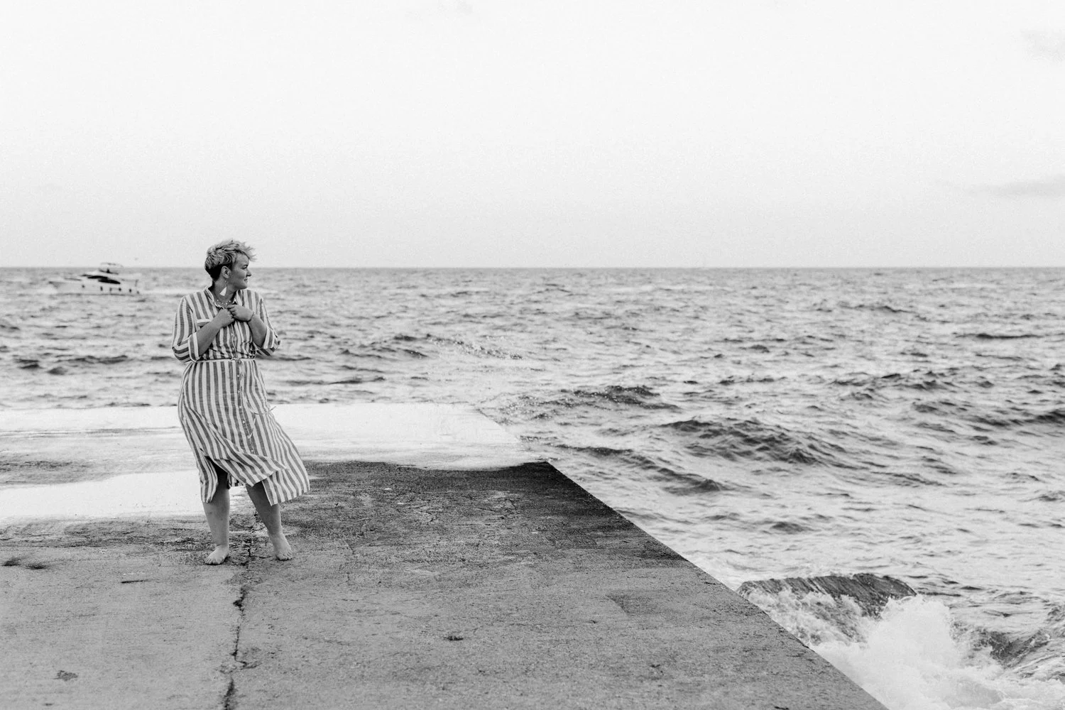Frau in gestreiftem Kleid steht barfuß auf einer Mole am Meer, blickt nach links, während die Wellen gegen das Ufer schlagen, schwarz-weiß Foto.