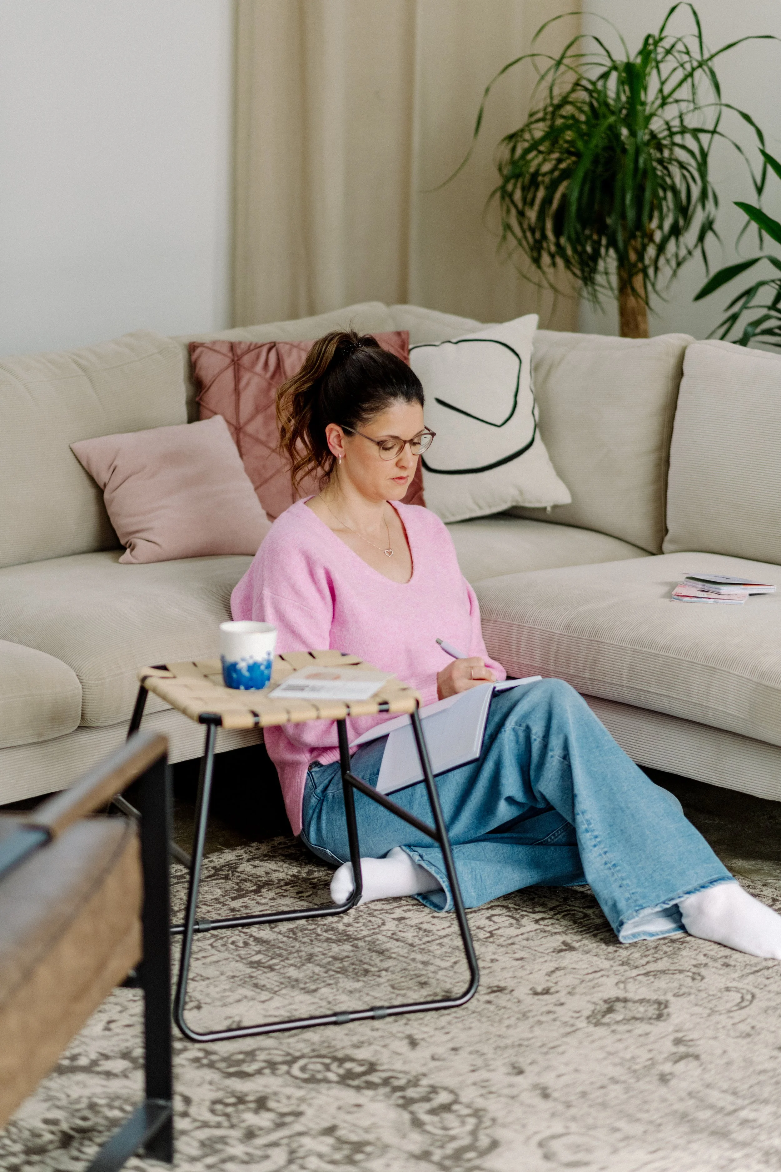 Eine Frau sitzt auf dem Boden vor einem Sofa, schreibt in ein Notizbuch und trinkt aus einer Tasse, während sie entspannt in einem Wohnzimmer ist.