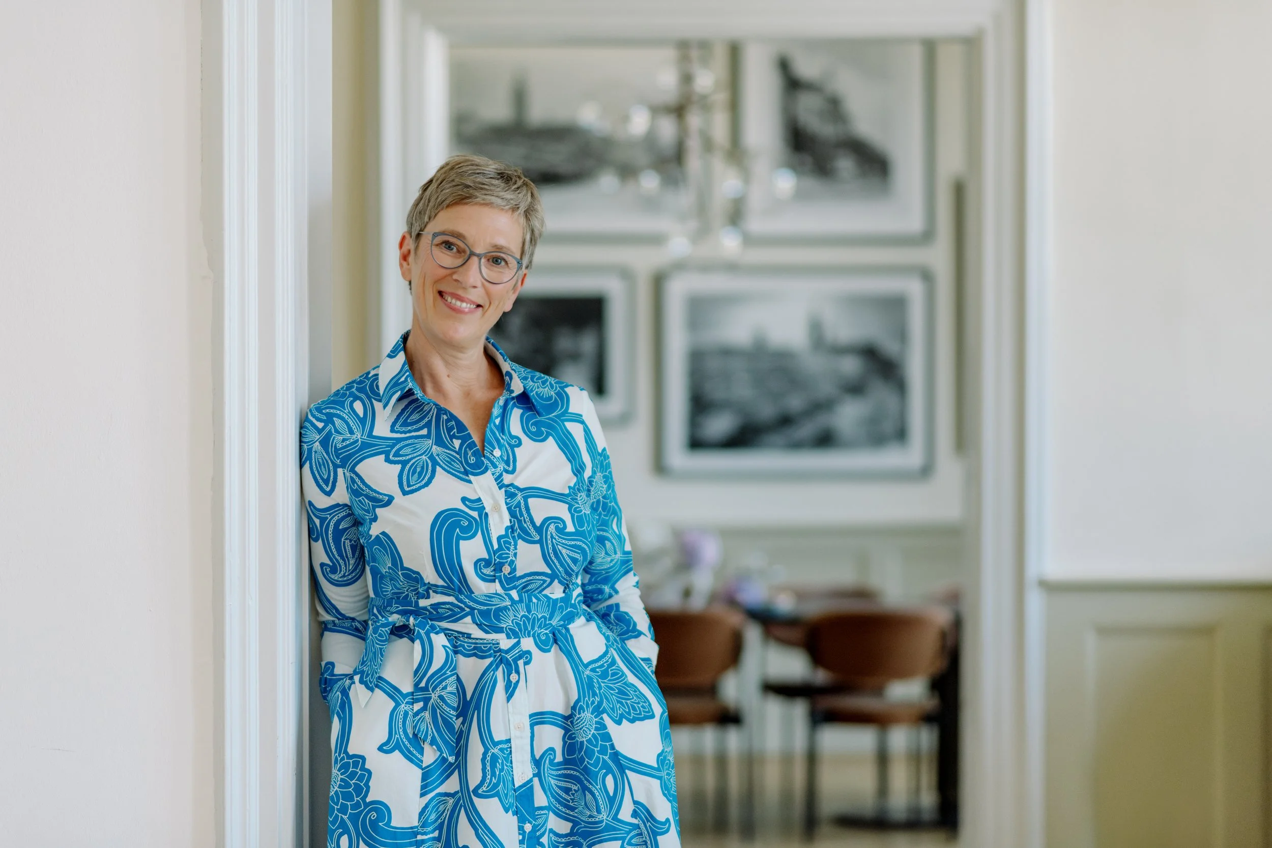 Eine lachende Frau mit kurz grauhaarigem Haar und Brille steht in einem weißen Hausflur, getragen eines blauen und weißen Kleid mit Blumenmuster, im Hintergrund sind mehrere Bilder an der Wand zu sehen.