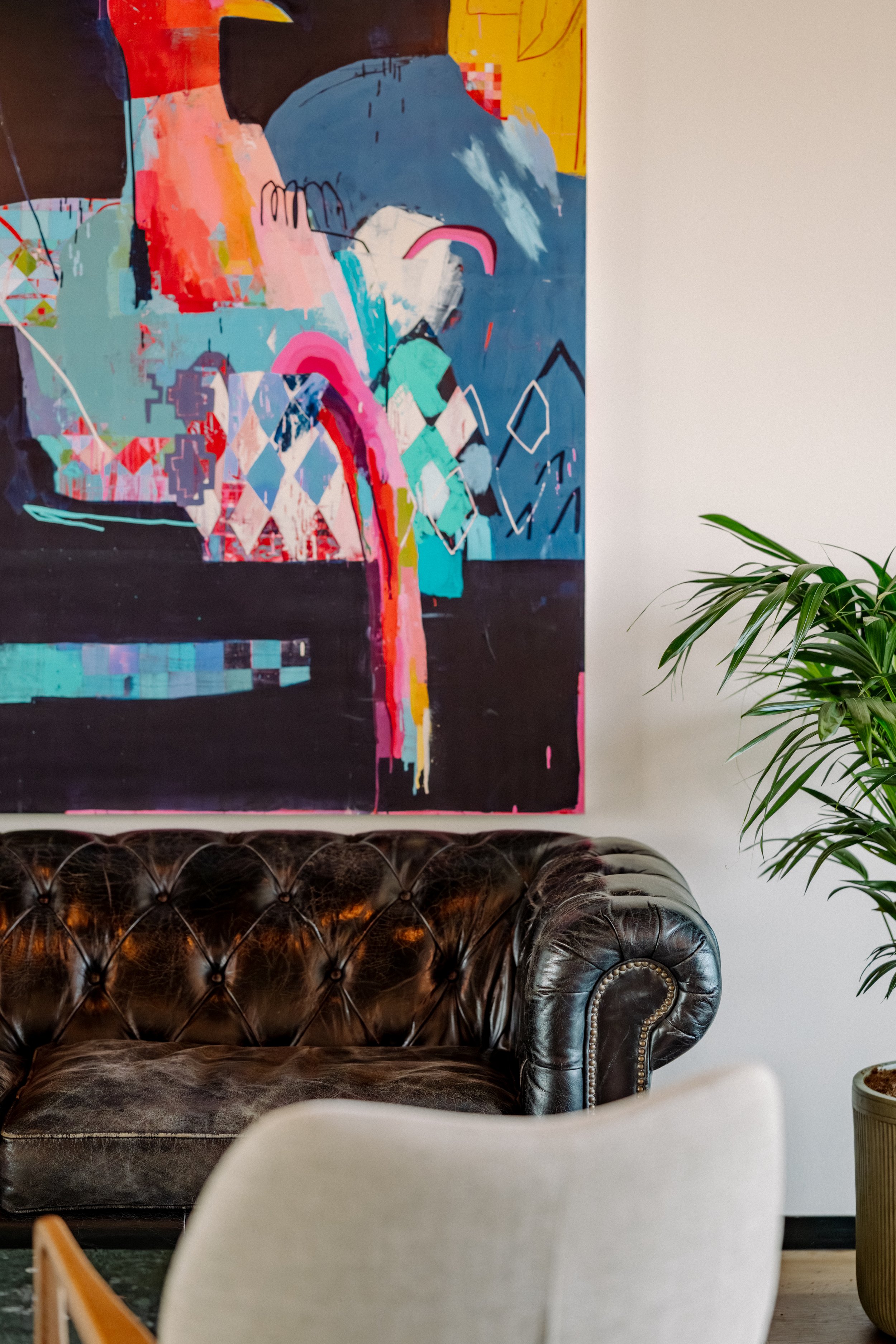 Ein braunes Ledersofa mit Knopf-Polsterung im Vordergrund, eine weiße Sitzgelegenheit, eine bunte abstrakte Wandkunst im Hintergrund und eine grüne Zimmerpflanze auf der rechten Seite.