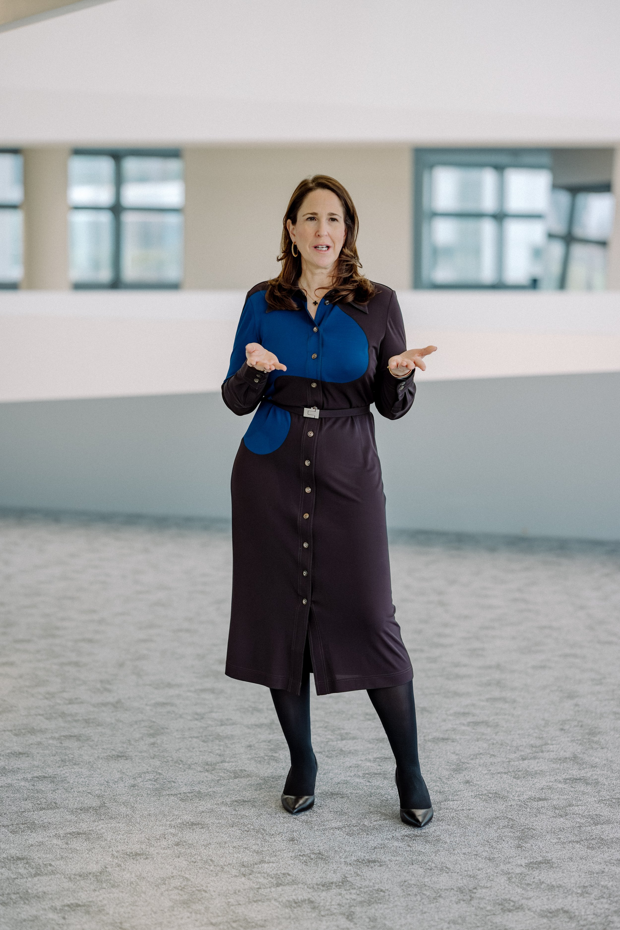 Eine Frau steht auf einem grauen, leeren Platz mit modernen Gebäuden im Hintergrund, trägt ein langes schwarzes Kleid mit blauen Akzenten, schwarze Strümpfe und High Heels, und hat braune, lockige Haare.