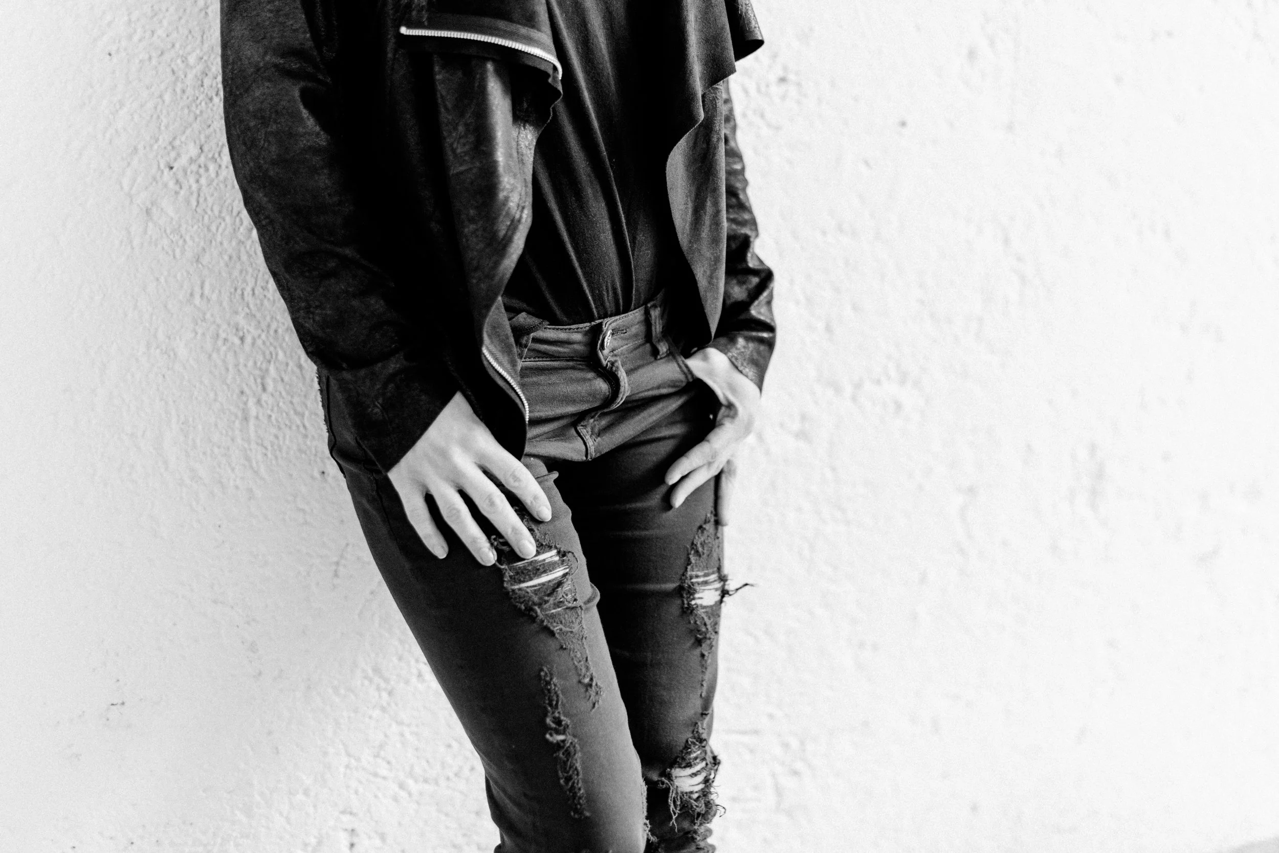 Nahaufnahme einer Person, die eine schwarze Lederjacke, Jeans mit Rissen und eine schwarze Hose trägt, vor einer weißen Wand steht.