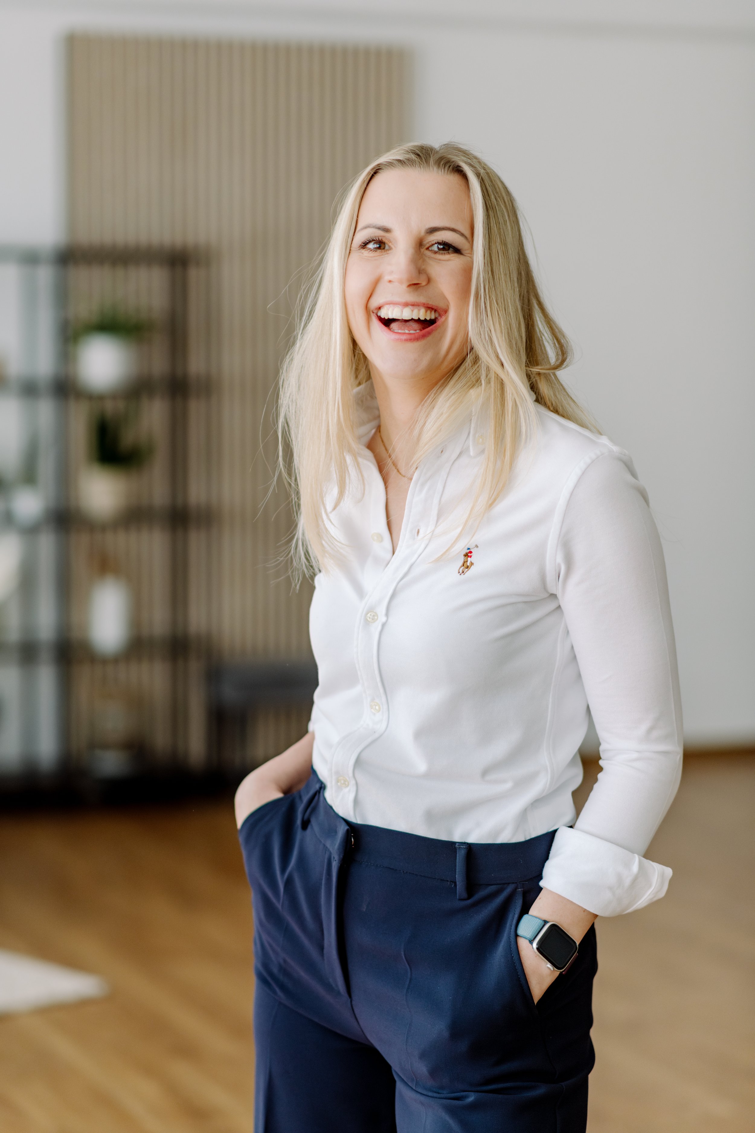 Lächelnde blonde Frau in Weißer Bluse und dunklen Hosen, stehend in einem modernen, leicht eingerichteten Raum mit Holzfußboden.