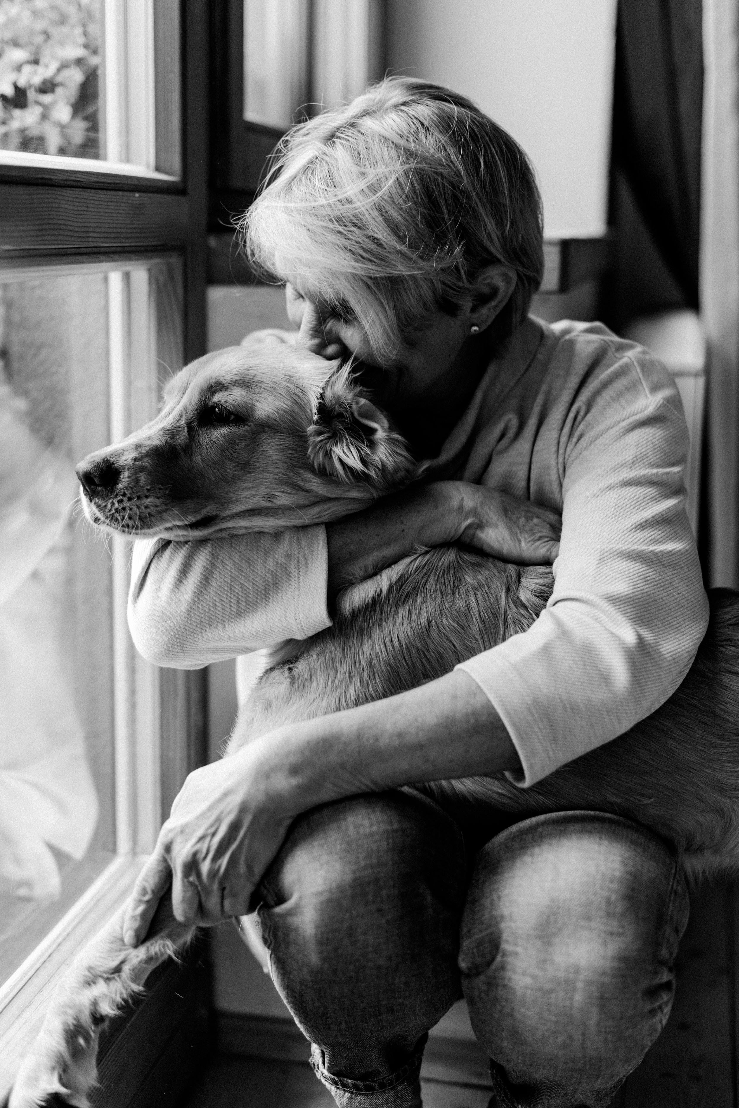 Eine ältere Frau mit kurzen Haaren und einem Ohrstecker, die einen Golden Retriever an einem Fenster umarmt, das in Schwarzweiß aufgenommen wurde.