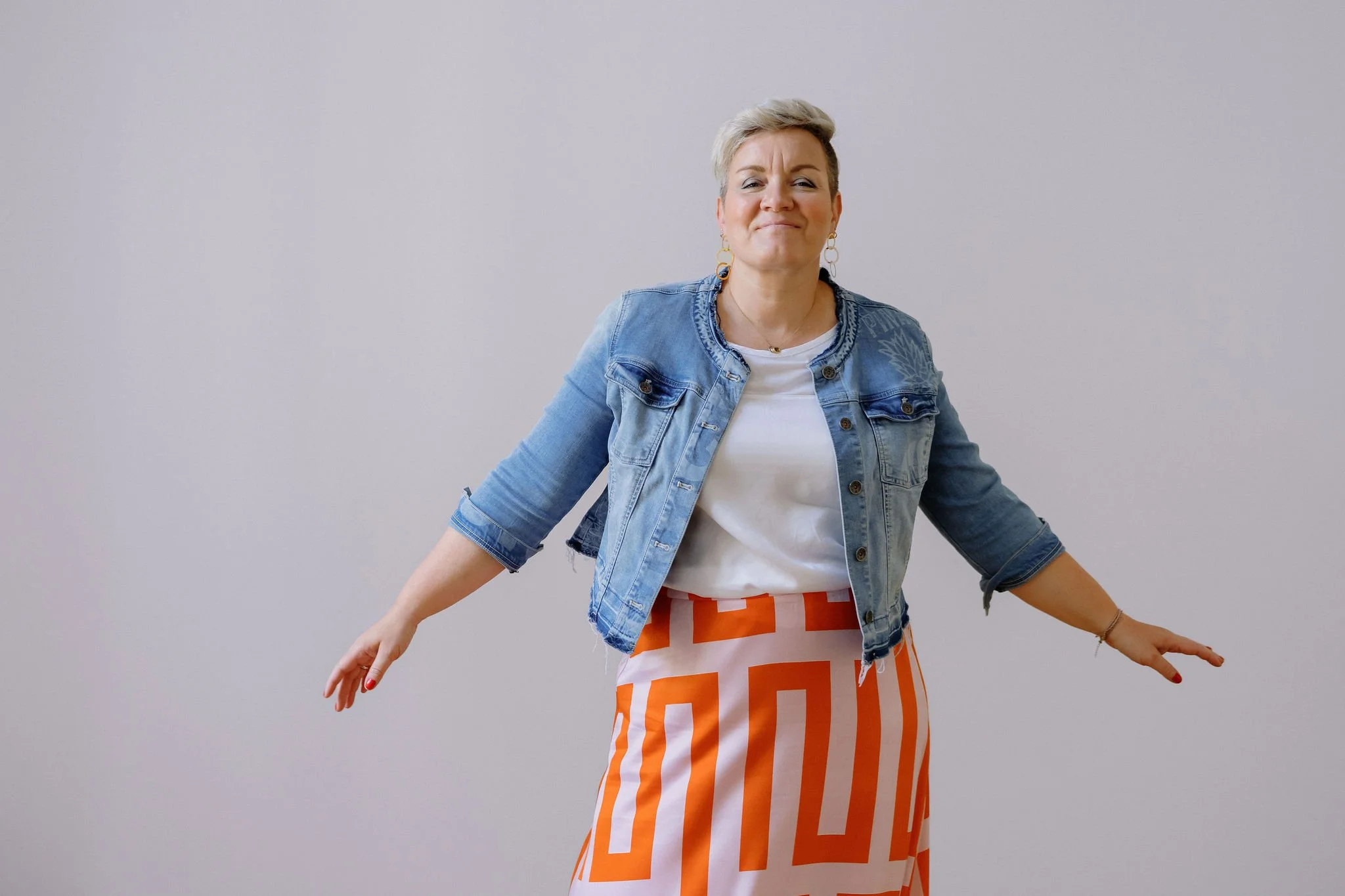 Eine Frau mit kurzen, blonden Haaren trägt eine Jeansjacke, eine weiße Bluse und eine orange-weiße gestreifte Rock. Sie steht vor einer hellgrauen Wand, ihre Arme sind leicht ausgestreckt, und sie lächelt leicht.