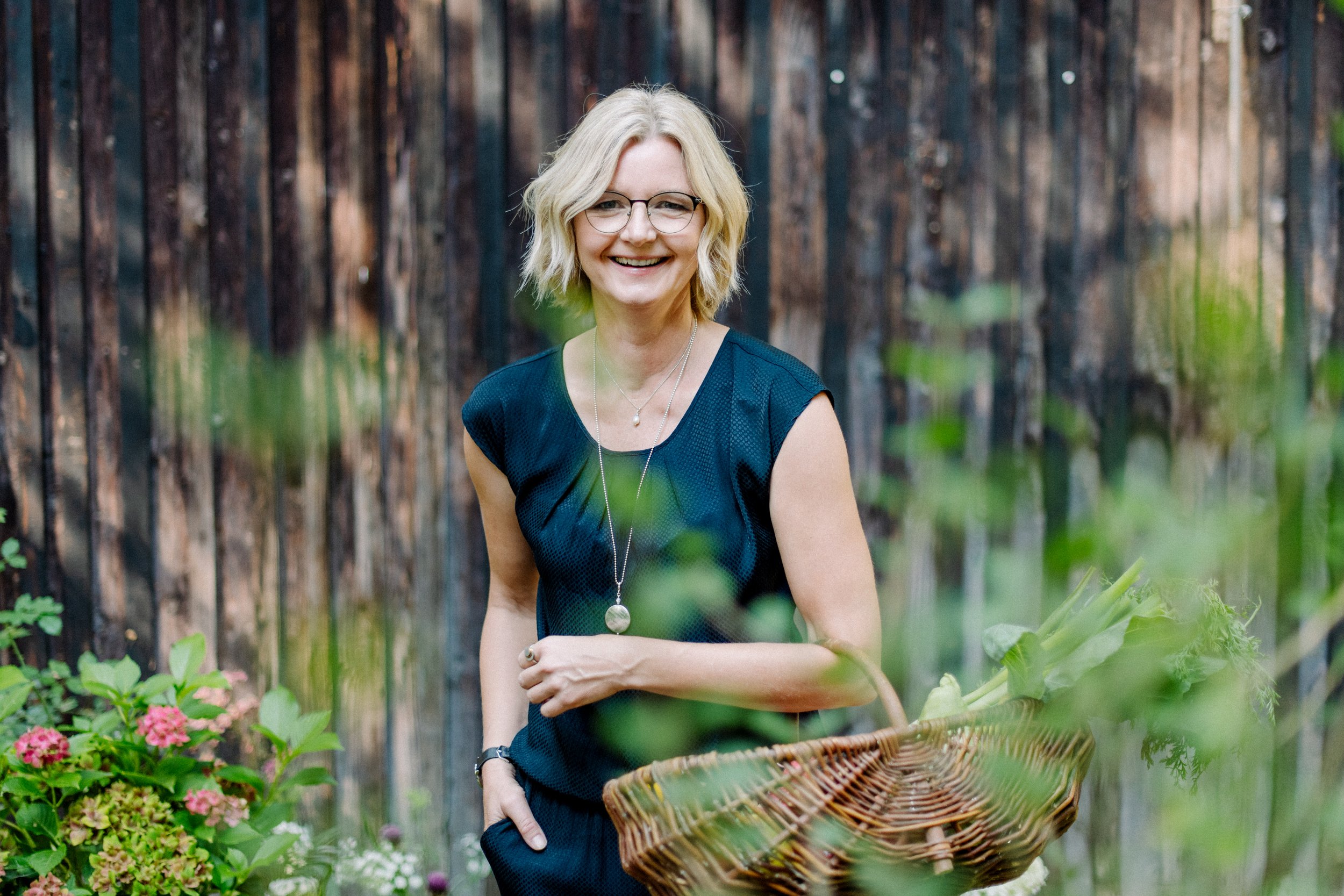 Eine lachende Frau mit blonden Haaren, Brille und schwarzem Oberteil, die einen Korb mit Blumen in einem Garten hält.