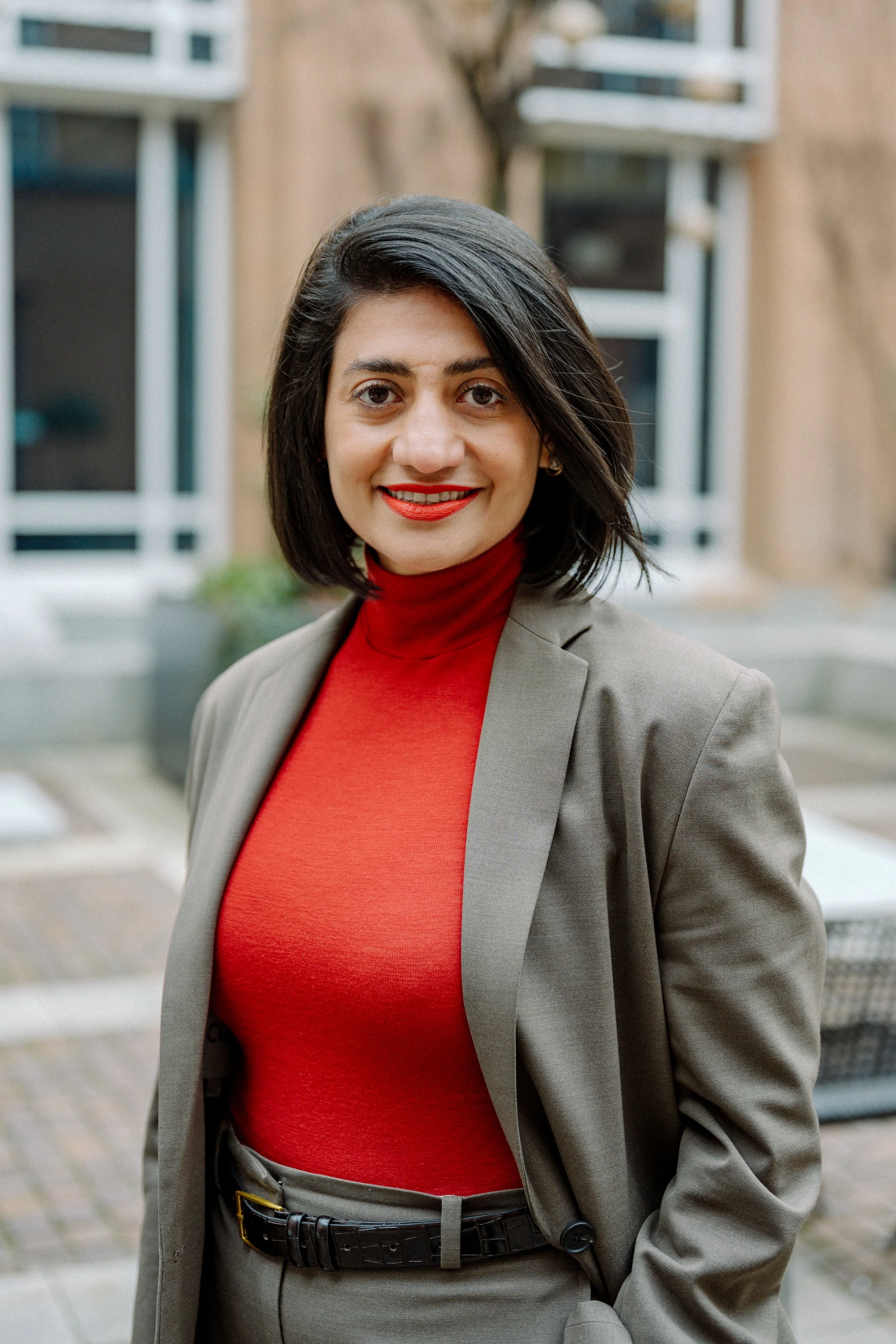Porträt einer Frau mit schwarzem Haar, rotem Pullover, grauem Blazer und roter Lippenstift vor einem urbanen Hintergrund.