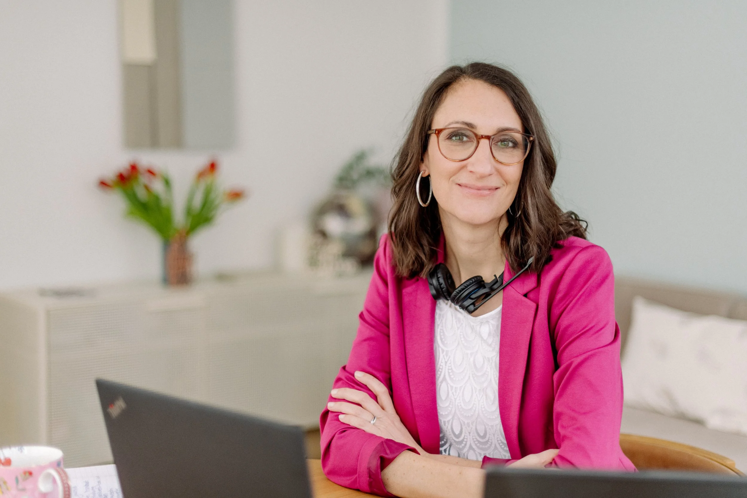 Eine Frau mit braunen, lockigen Haaren, Brille und Ohrringen sitzt an einem Tisch in einem Büro. Sie trägt eine rosa Jacke und ein weißes Oberteil. Im Hintergrund ist ein Spiegel und eine Vase mit roten Blumen sichtbar.