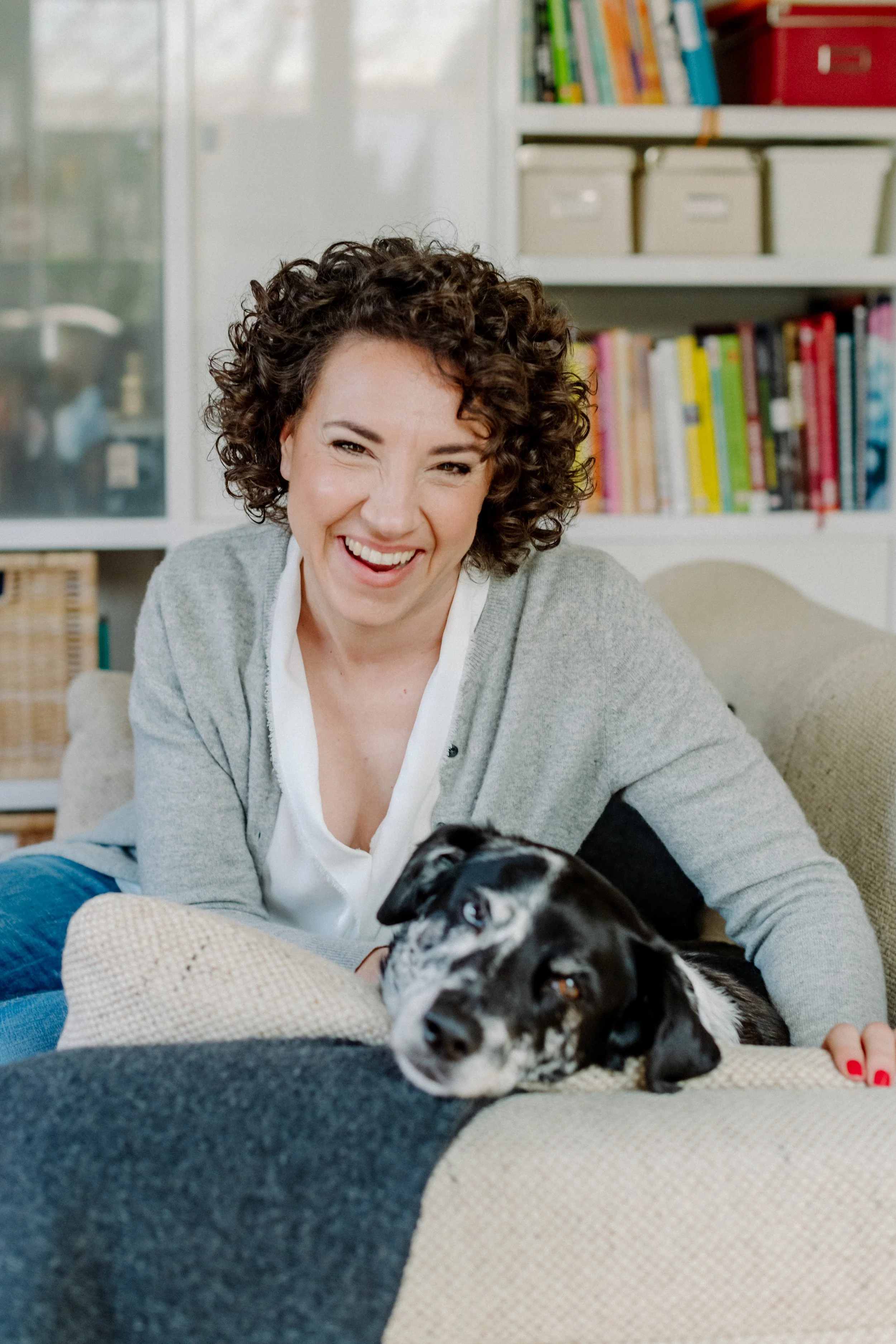 Lächelnde Frau mit dunkelbraunen, lockigen Haaren, die auf einem Sofa sitzt, mit einem blauen und einem beigen Kissen, neben einem schwarzen und weißen Hund, im Hintergrund ein Bücherregal.