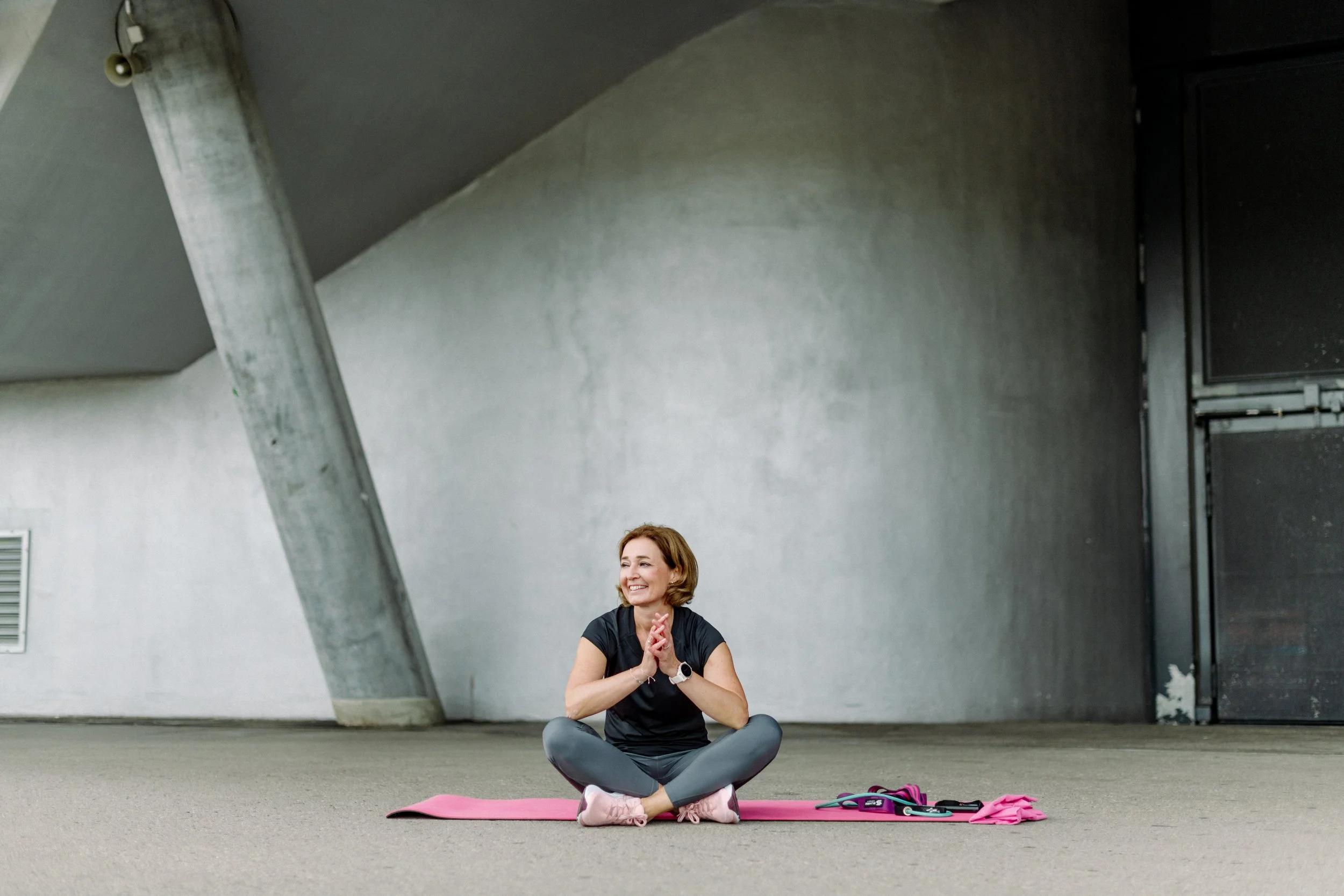 Eine Frau sitzt auf einer Yogamatte im Freien vor einer Betonwand, lächelt und wirkt entspannt. Sie trägt sportliche Kleidung, eine Armbanduhr und befindet sich vor einem grauen Hintergrund mit einer Metallstütze und einer Tür.