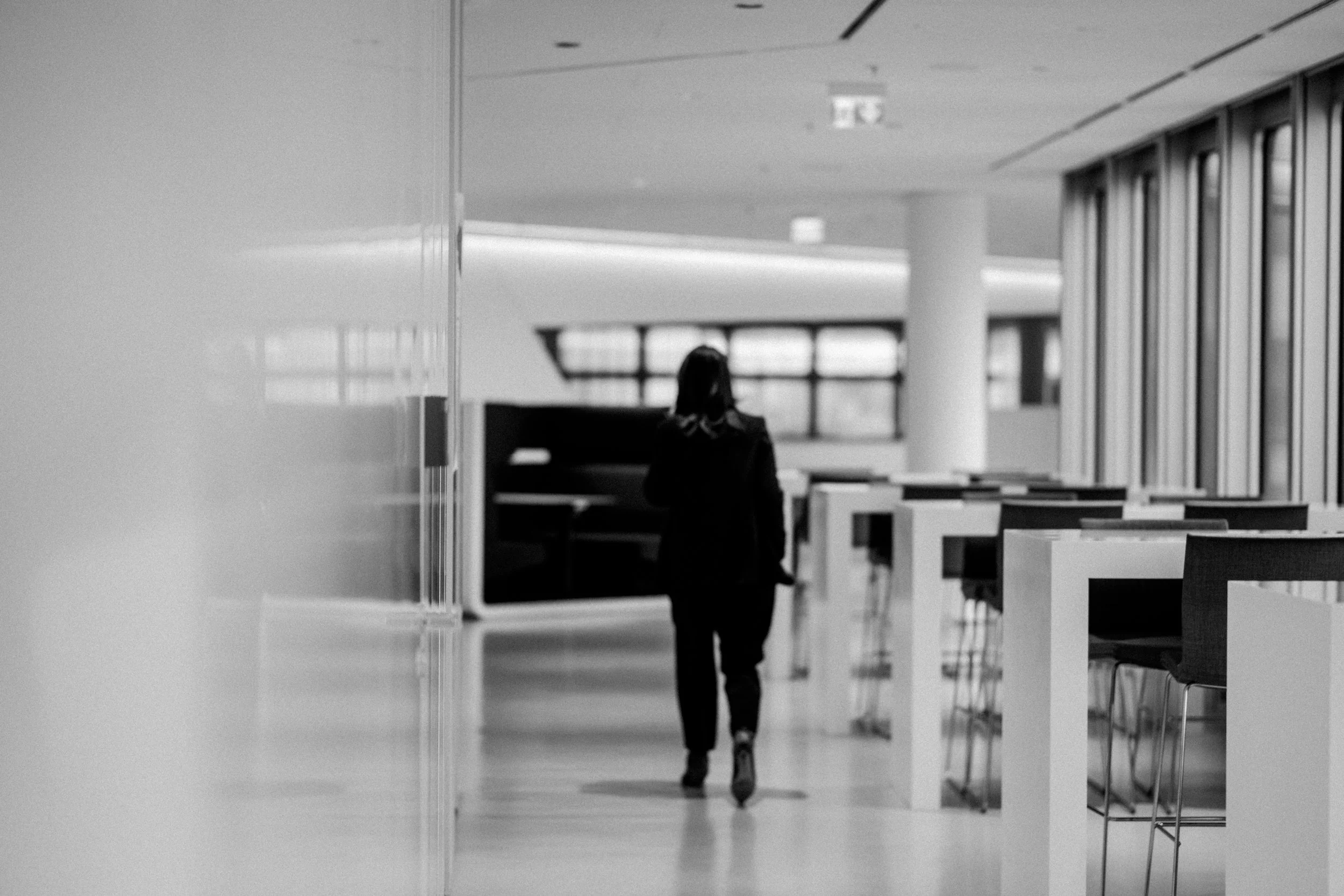 Eine Person, vermutlich eine Frau, läuft in einem modernen, hell erleuchteten Innenraum, möglicherweise einem Flughafen oder Bahnhof. Der Raum ist mit Tischen und Stühlen ausgestattet und hat große Fenster, die Tageslicht hereinscheinen lassen.