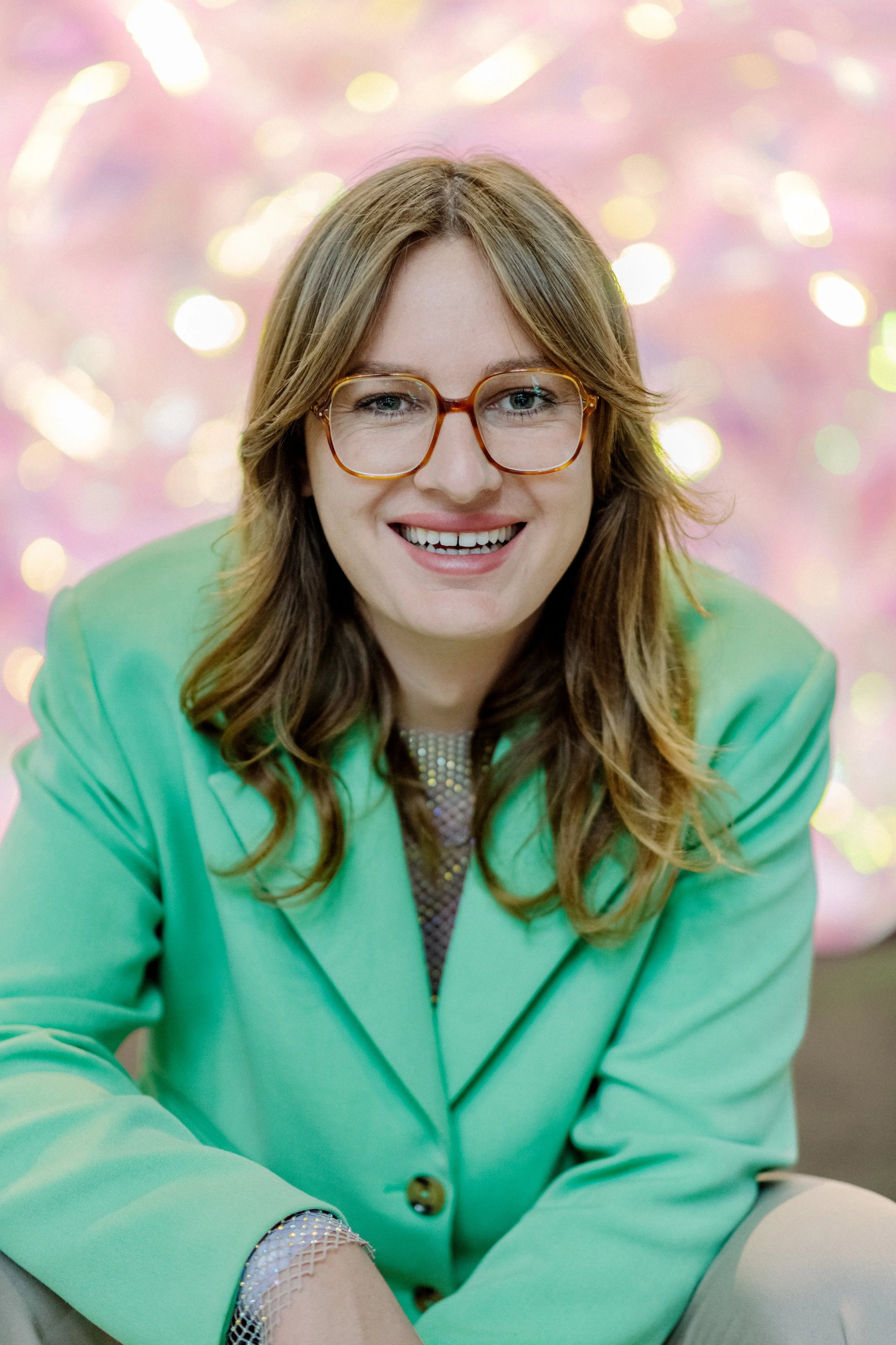 Jugendliche Frau mit braunen Locken, braunen Brille und hellem Teint, lachend, trägt eine grüne Jacke, im Hintergrund bunte Lichter im Bokeh-Effekt.
