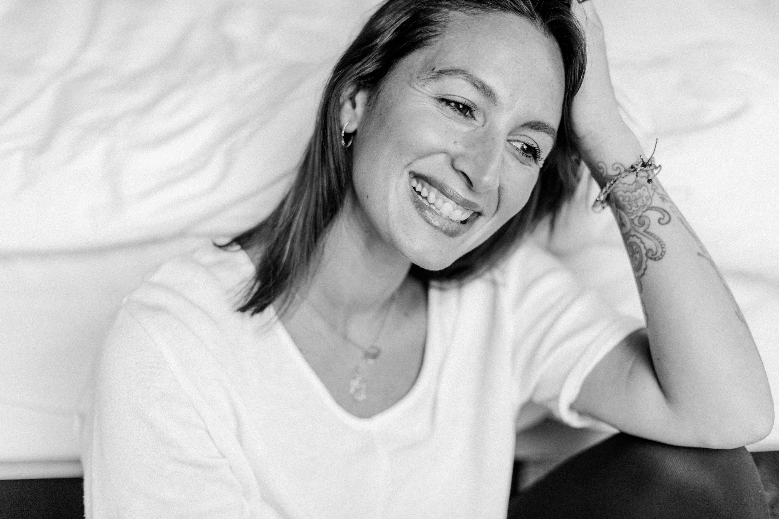 Lächelnde junge Frau mit dunklen Haaren und Tattoos auf dem Arm, entspannt sitzend vor einem weißem Bett, in Schwarzweiß fotografiert.
