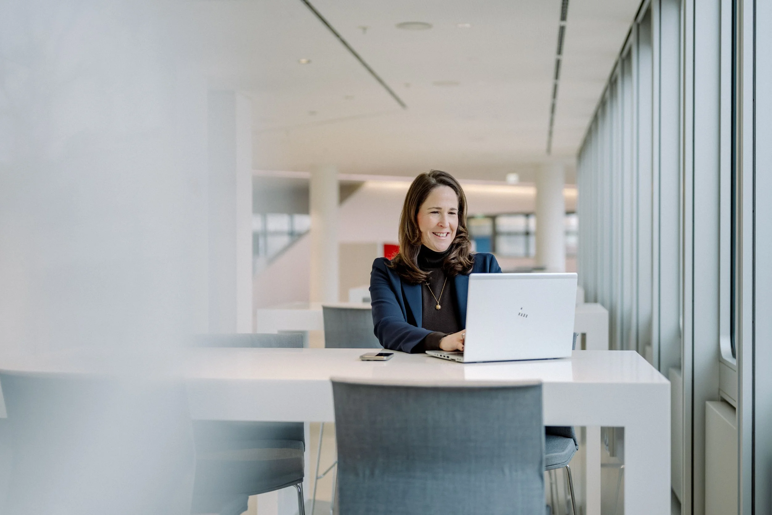 Eine lachende Frau sitzt an einem weißen Tisch in einem modernen, hell erleuchteten Büro und arbeitet an einem Laptop.