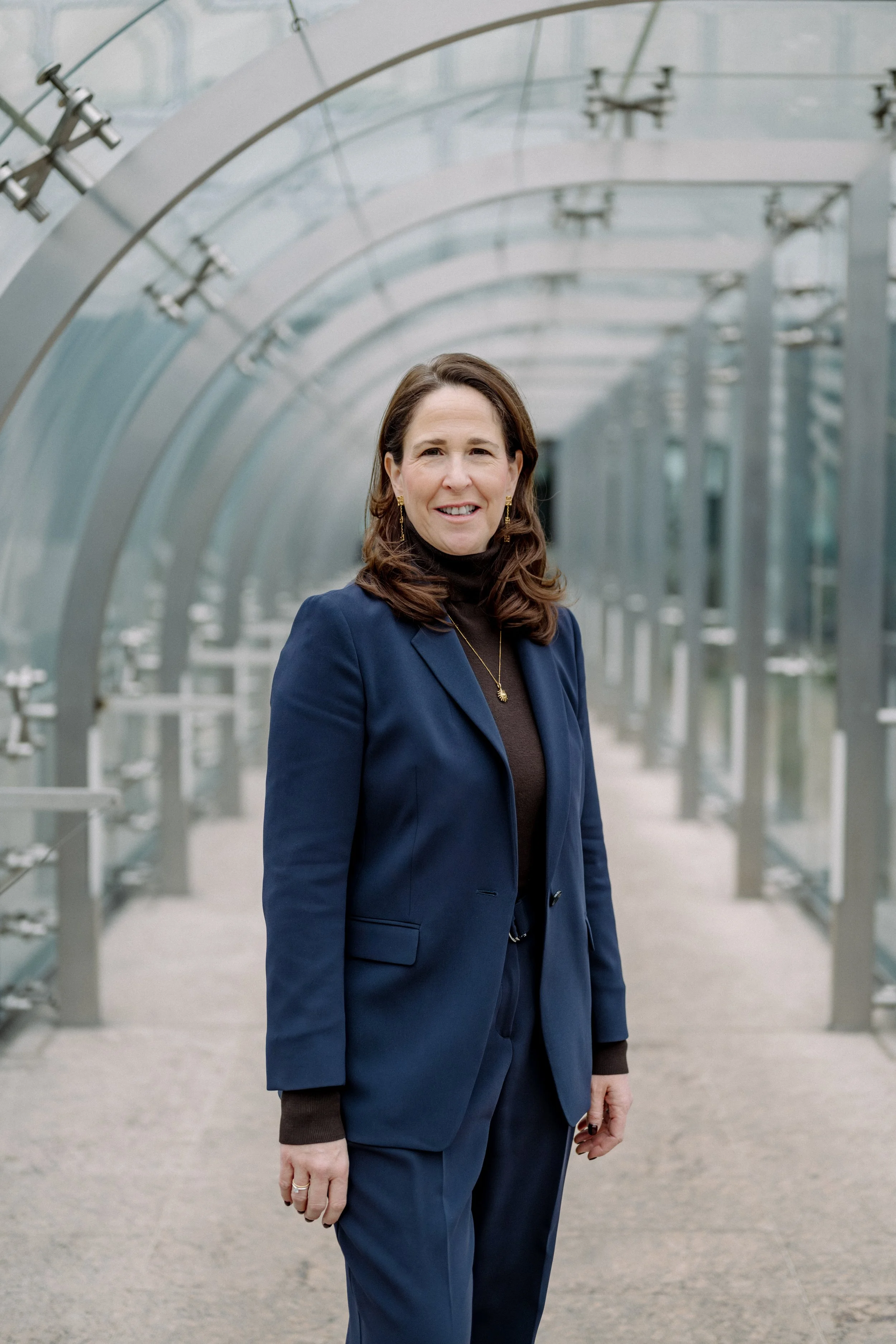Eine Frau trägt einen blauen Anzug und steht in einem Glashallen-Architektur. Sie lächelt leicht und trägt goldene Ohrringe sowie eine Halskette.