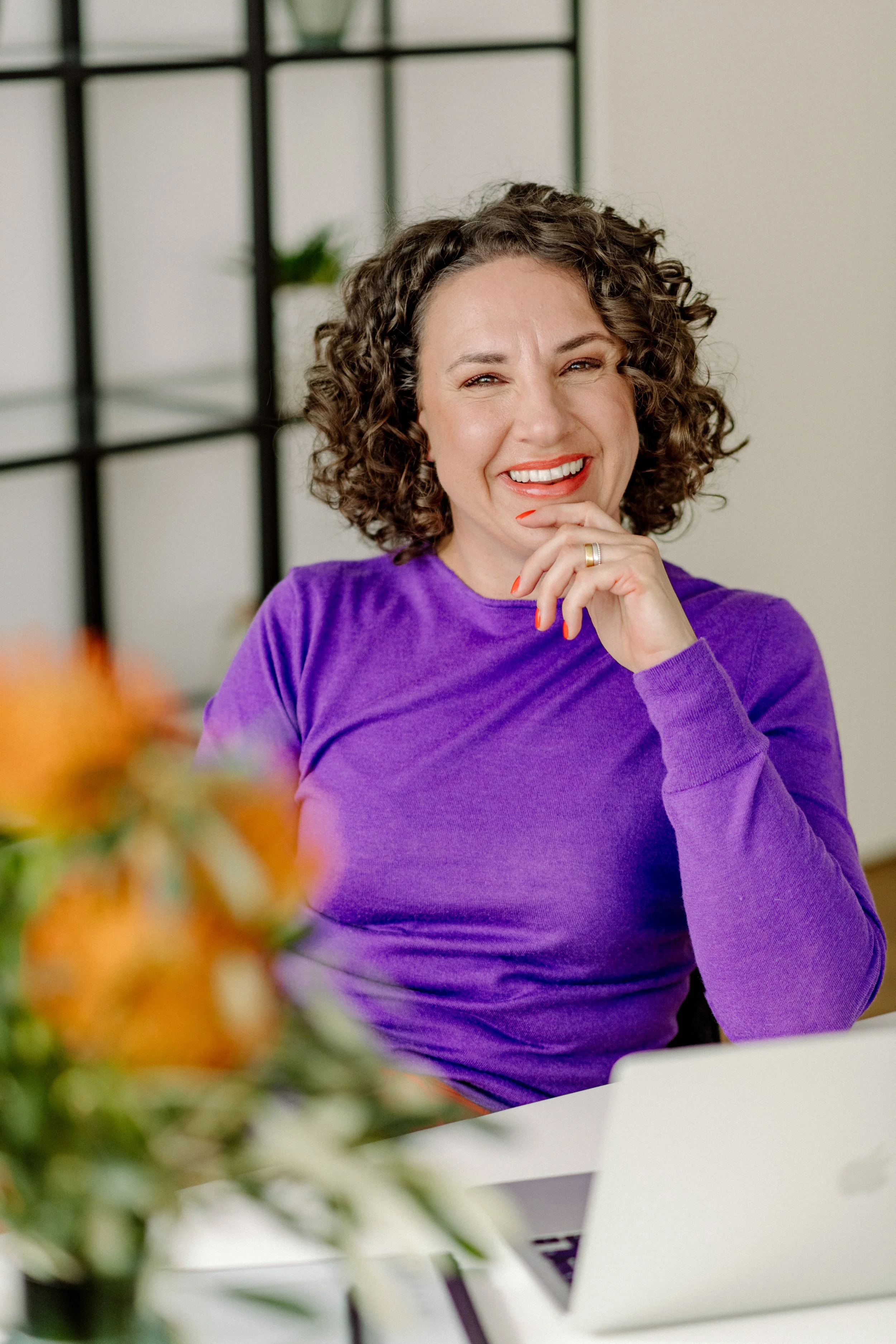 Frau mit lockigem Haar und rotem Lippenstift, lächelnd und die Hand am Kinn, sitzt an einem Tisch mit einem Laptop, trägt einen lila Pullover, im Hintergrund ein Regal mit Pflanzen.