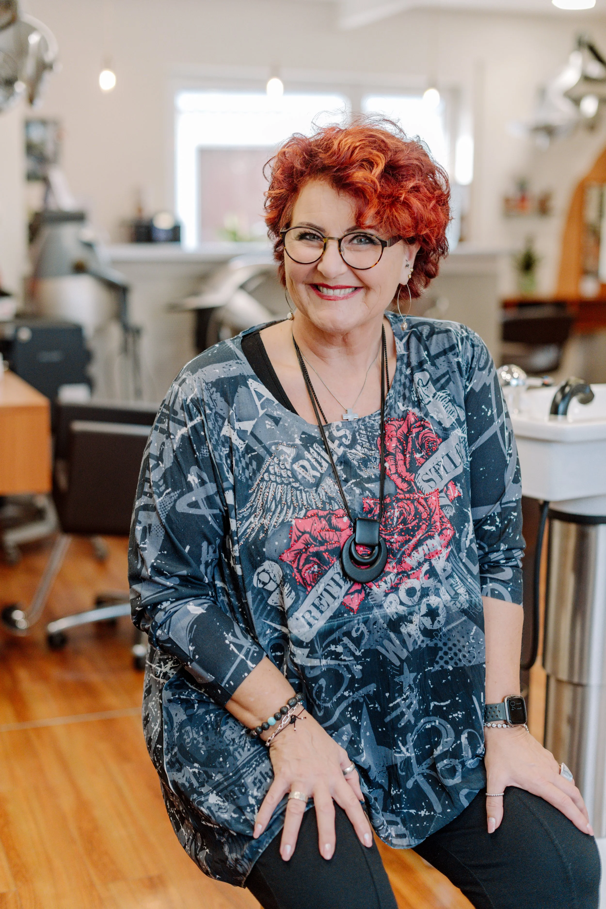 Ein lachender, rothaariger Frau mit Brille sitzt in einem Friseursalon mit Holzfußboden.