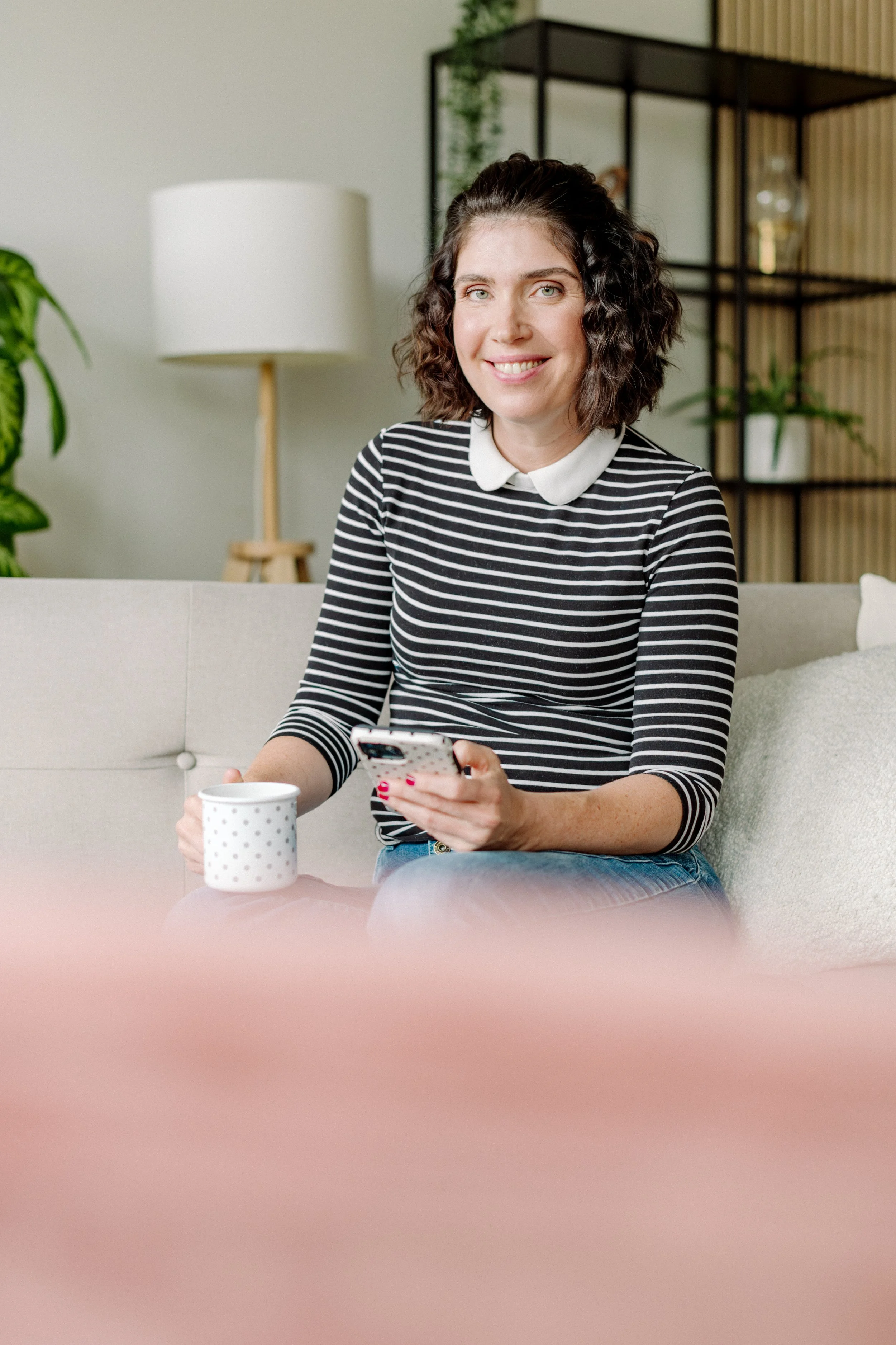 Eine lachende Frau mit kurzen, lockigen braunen Haaren sitzt auf einem Sofa, hält eine weiße Kaffeetasse mit Punkten in ihrer linken Hand und ein Smartphone in ihrer rechten Hand, während sie auf einem hellen, gemütlichen Wohnzimmer sitzt, mit Pflanz
