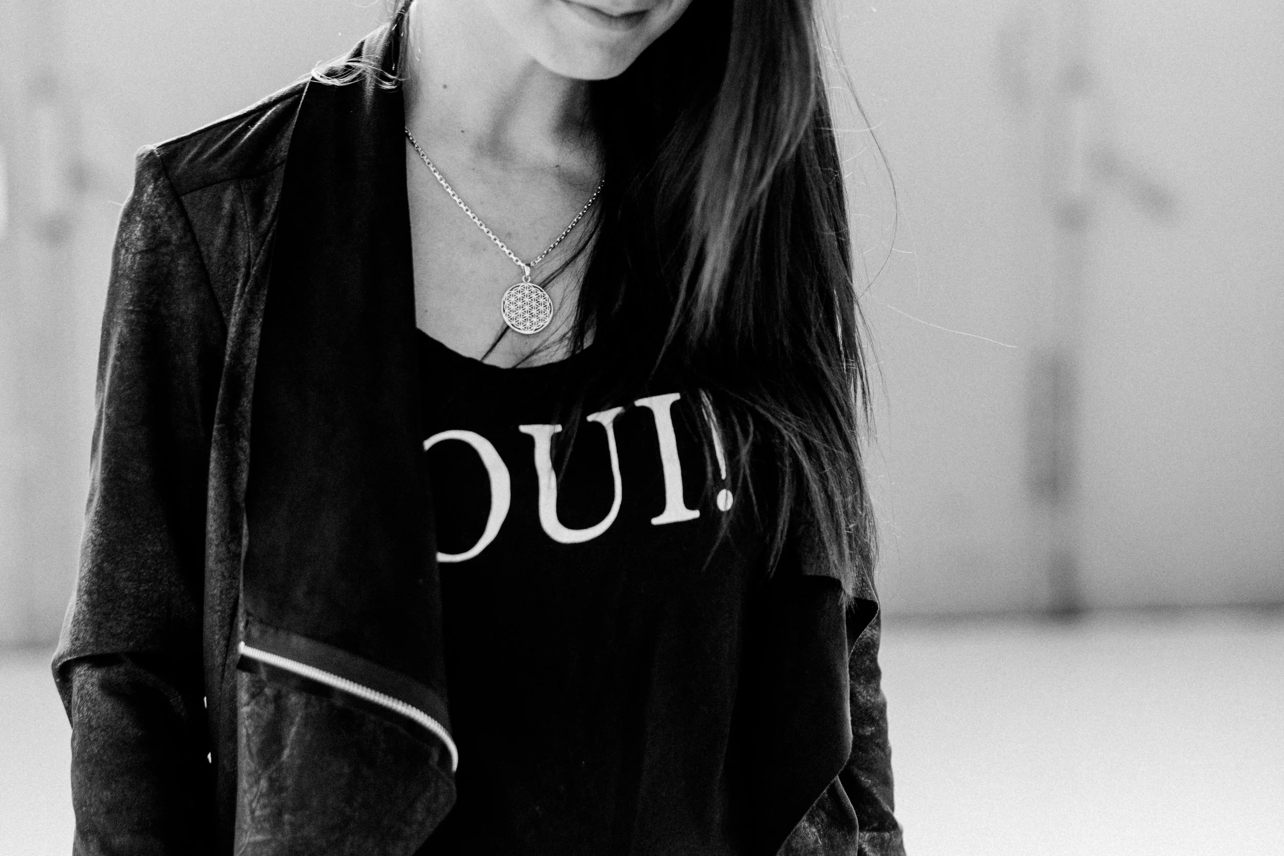 Nahaufnahme einer Frau, die nur den unteren Teil ihres Gesichts und ihren Oberkörper zeigt. Sie trägt eine Halskette und ein schwarzes T-Shirt mit weißen Buchstaben, sowie eine Jacke.