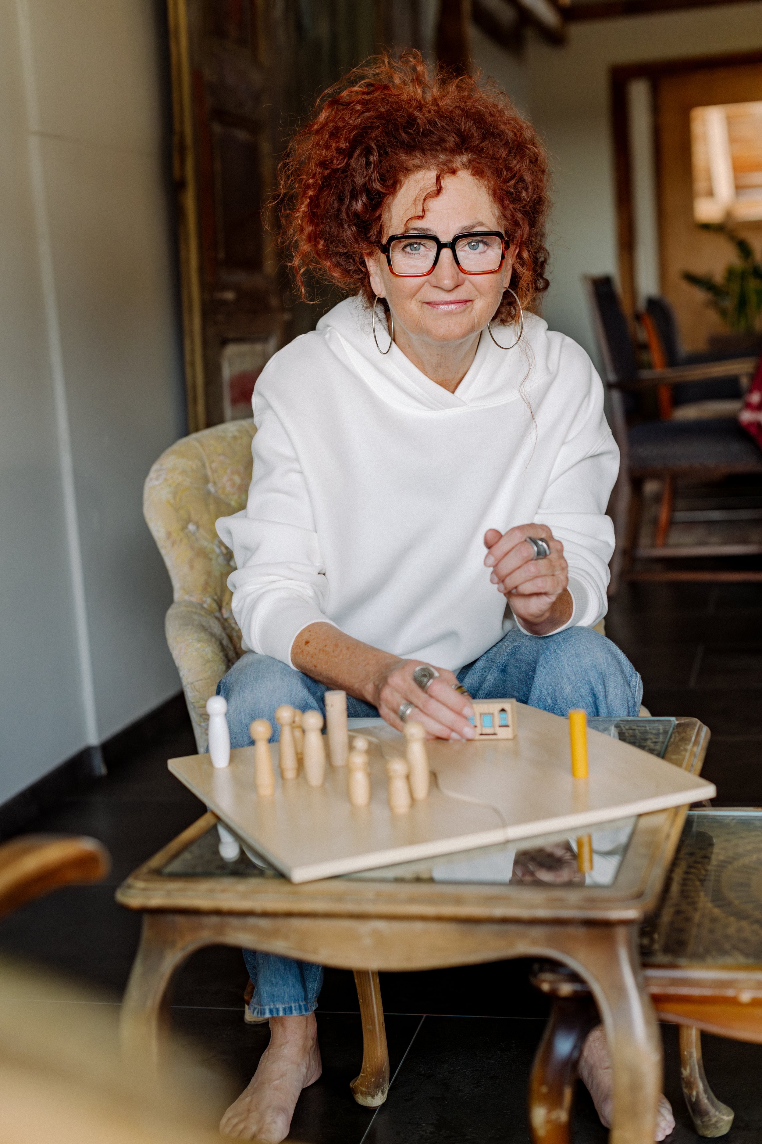 Eine ältere Frau mit roten, lockigen Haaren und Brille mit Aufstellungsfiguren, sitzend an einem Tisch.