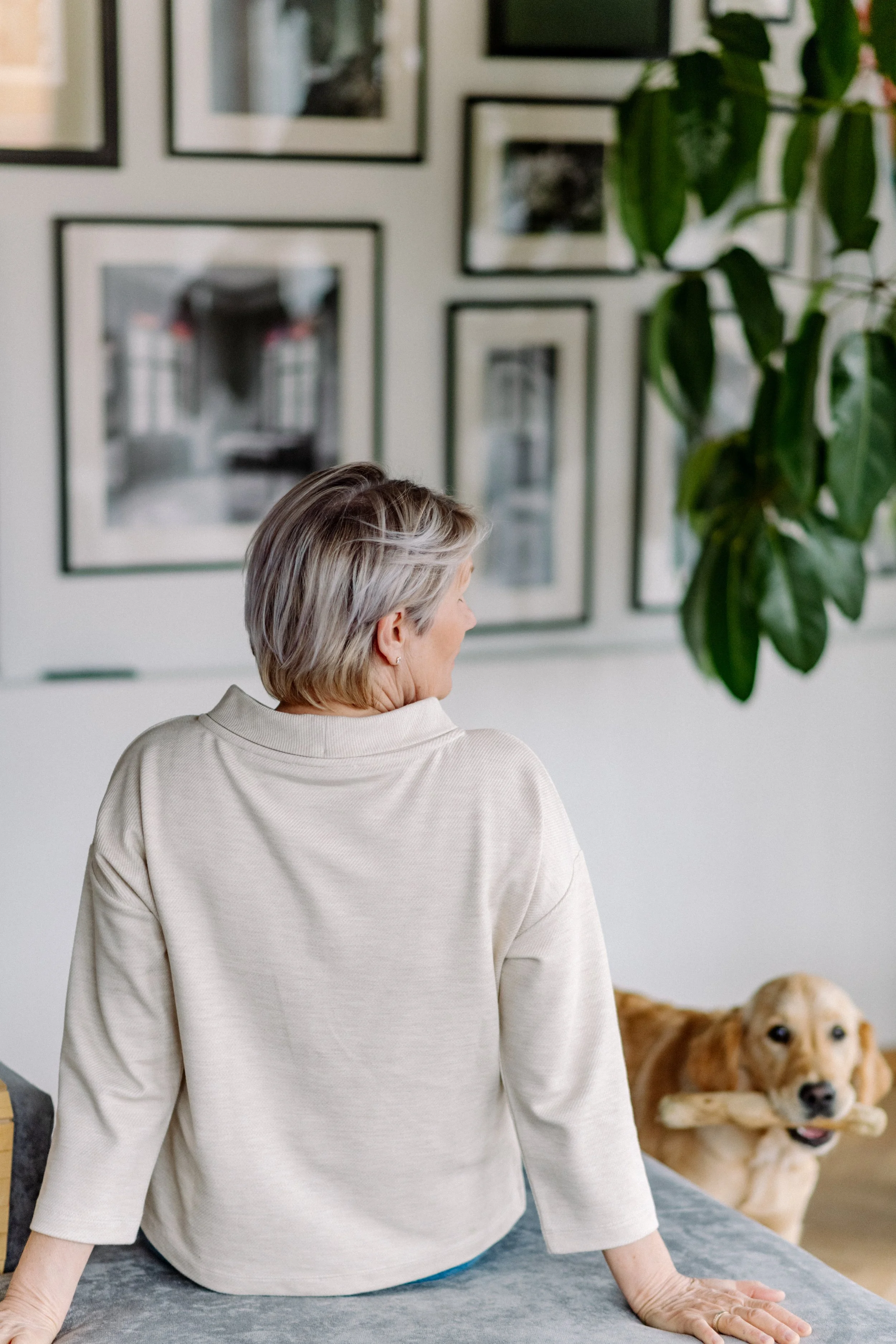Ältere Frau in beigem Pullover macht Yoga Position auf einem Teppich, hinter ihr ein Golden Retriever Hund sitzt auf dem Boden, im Hintergrund eine Wand mit mehreren Bilderrahmen und ein alter Baum.