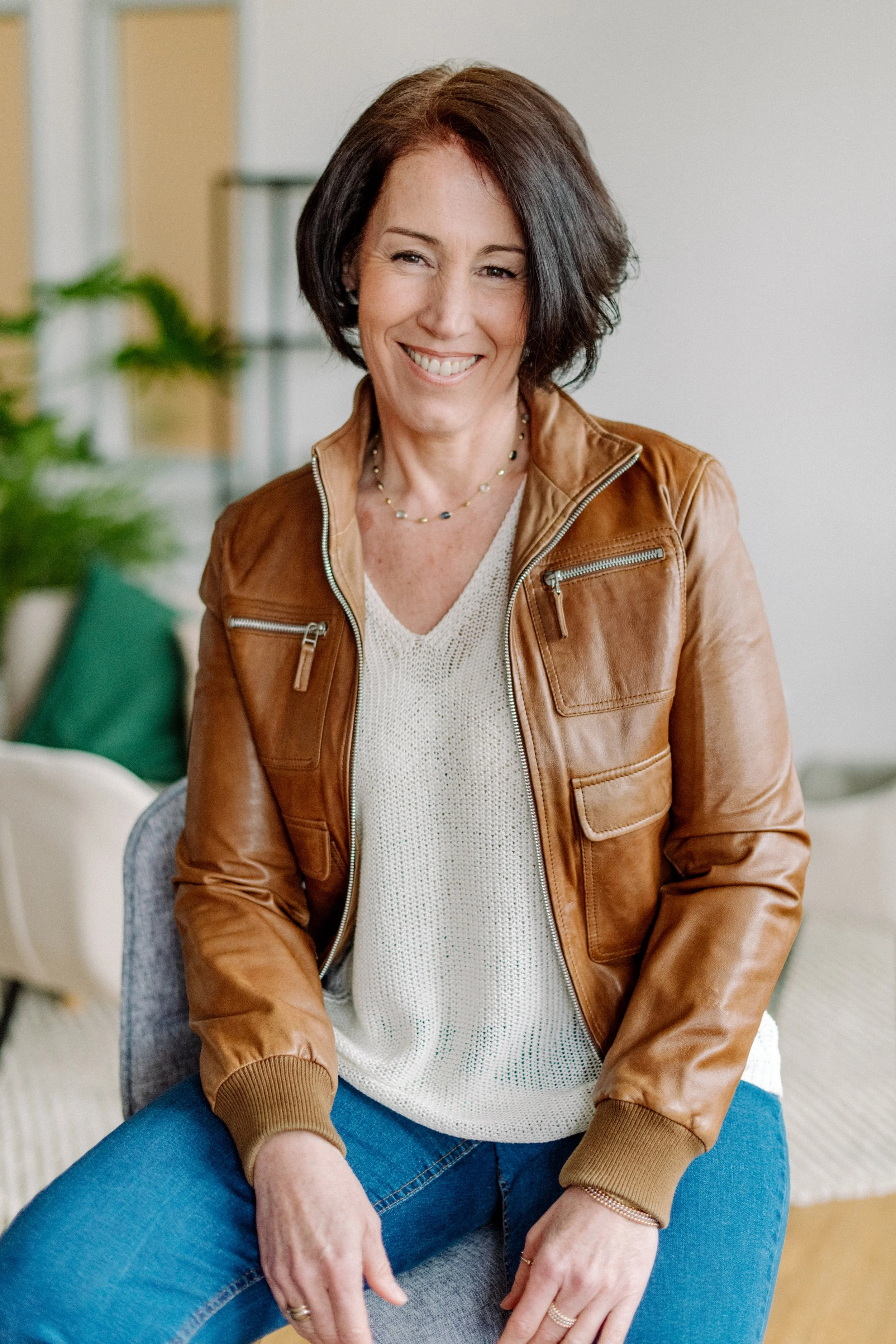 Frau mit brauner Lederjacke, weißen Pullover und blauen Jeans, lächelnd in einem modernen Raum.