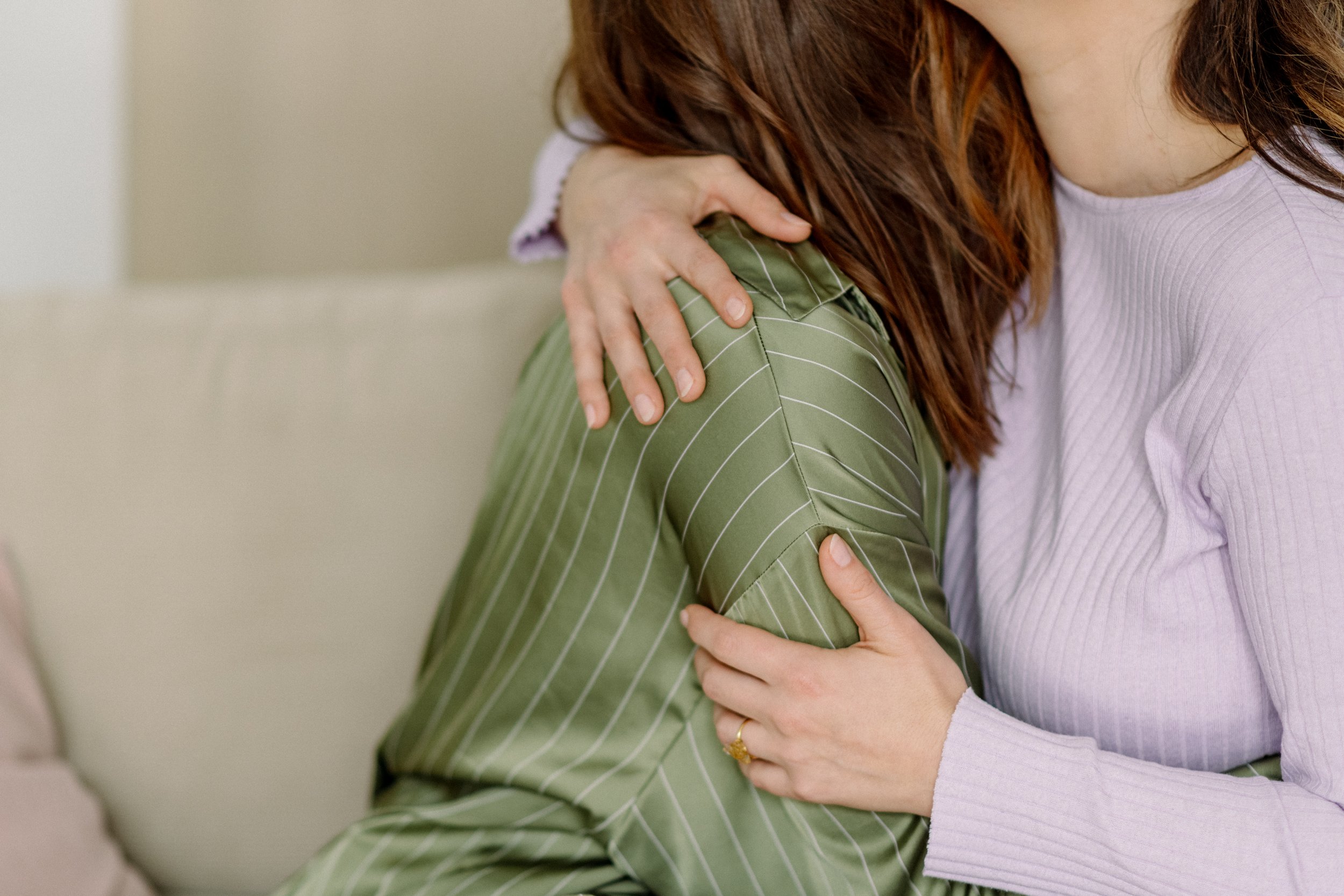 Zwei Frauen umarmen sich innig, eine trägt einen grünen karierten Mantel, die andere ein lilafarbenes Kleid. Der Hintergrund ist neutral gehalten.
