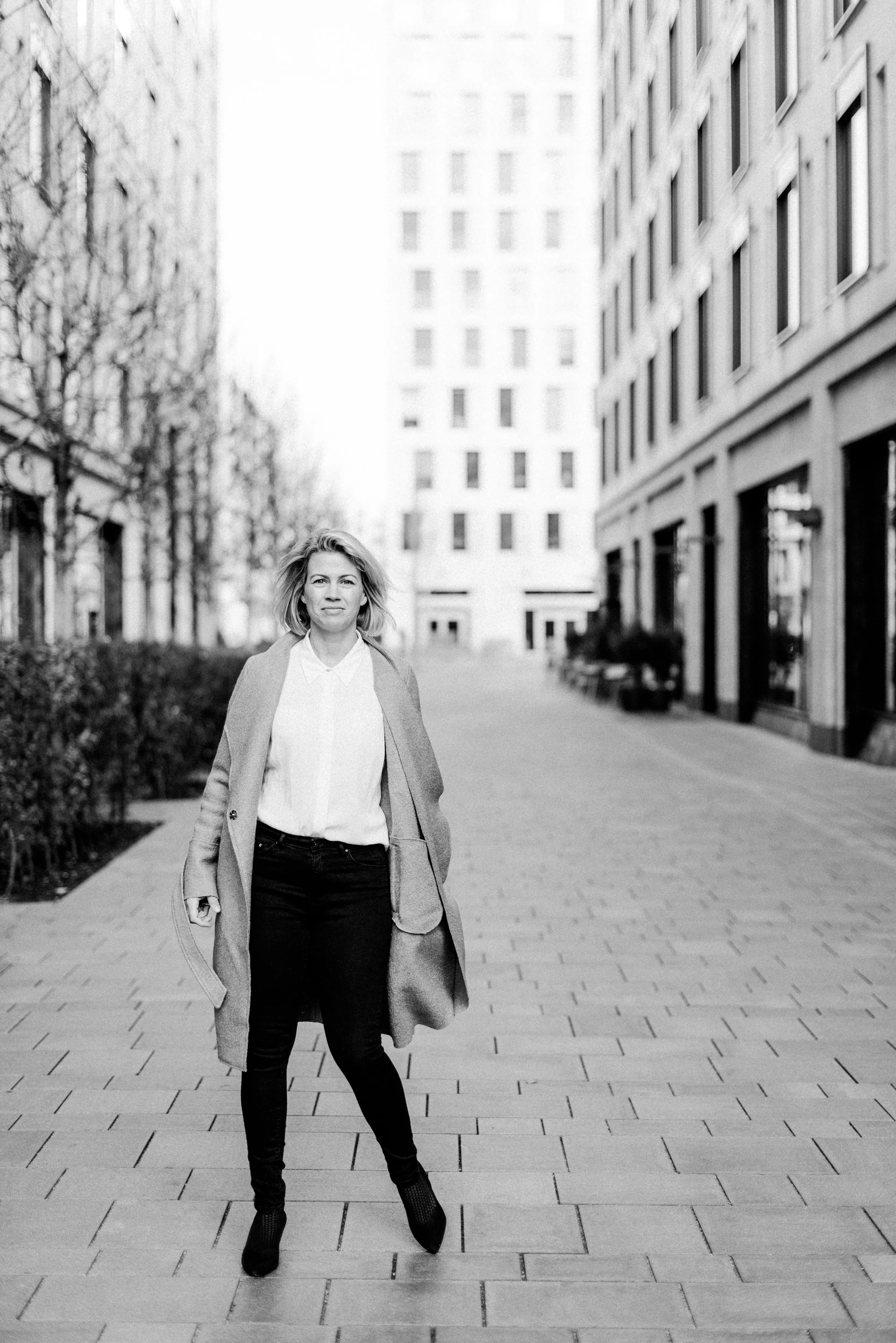Eine Frau in einem längeren Mantel, weißem Hemd, schwarzen Hosen und hohen Schuhen läuft auf einer gepflasterten Straße in einer städtischen Umgebung mit modernen Gebäuden und Bäumen.