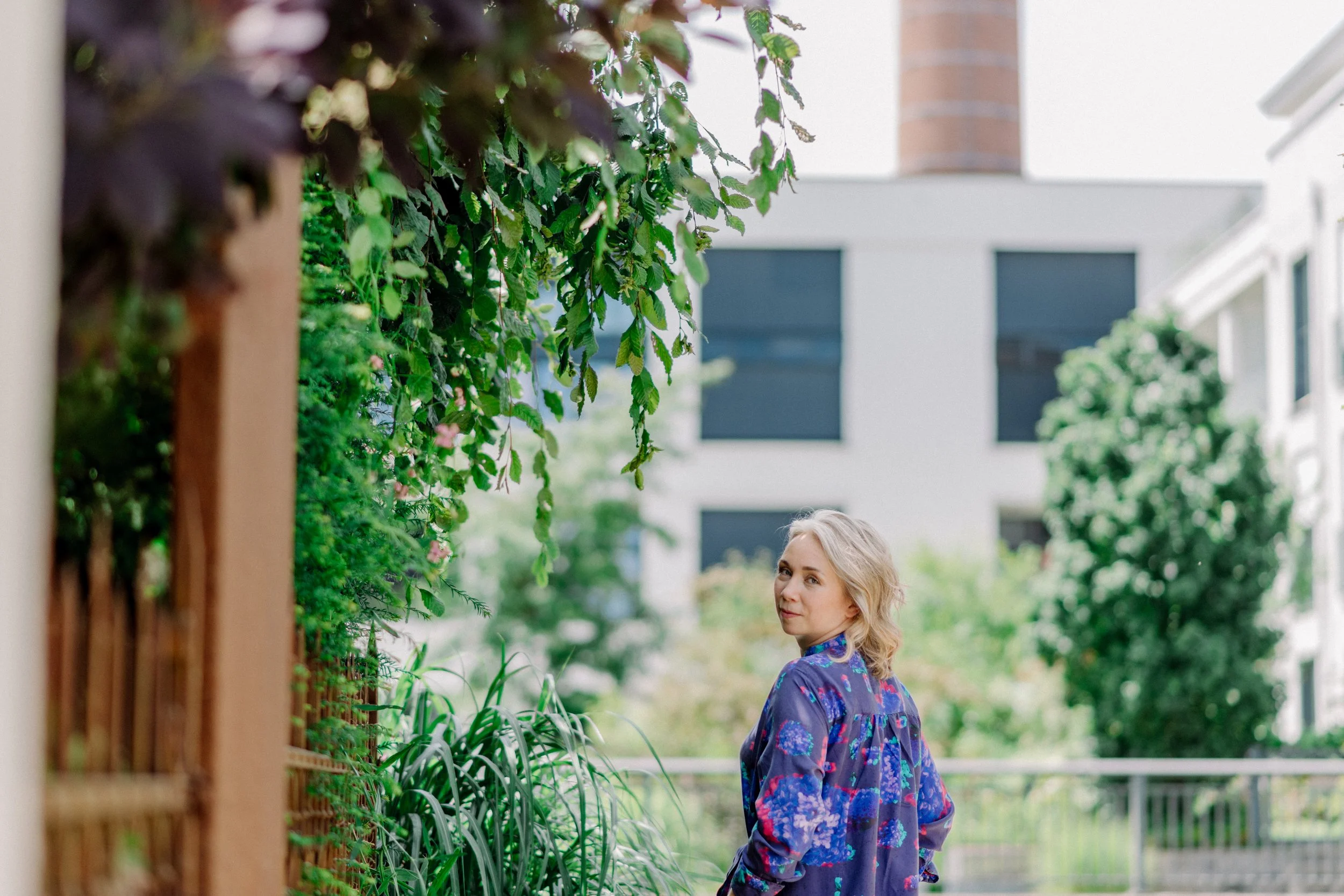 Junge Frau mit blauer, bunt gemusterter Bluse, stehend an einem grünen Gartenzaun, umgeben von Pflanzen, vor einem modernen weißen Haus mit dunklen Fensterläden.