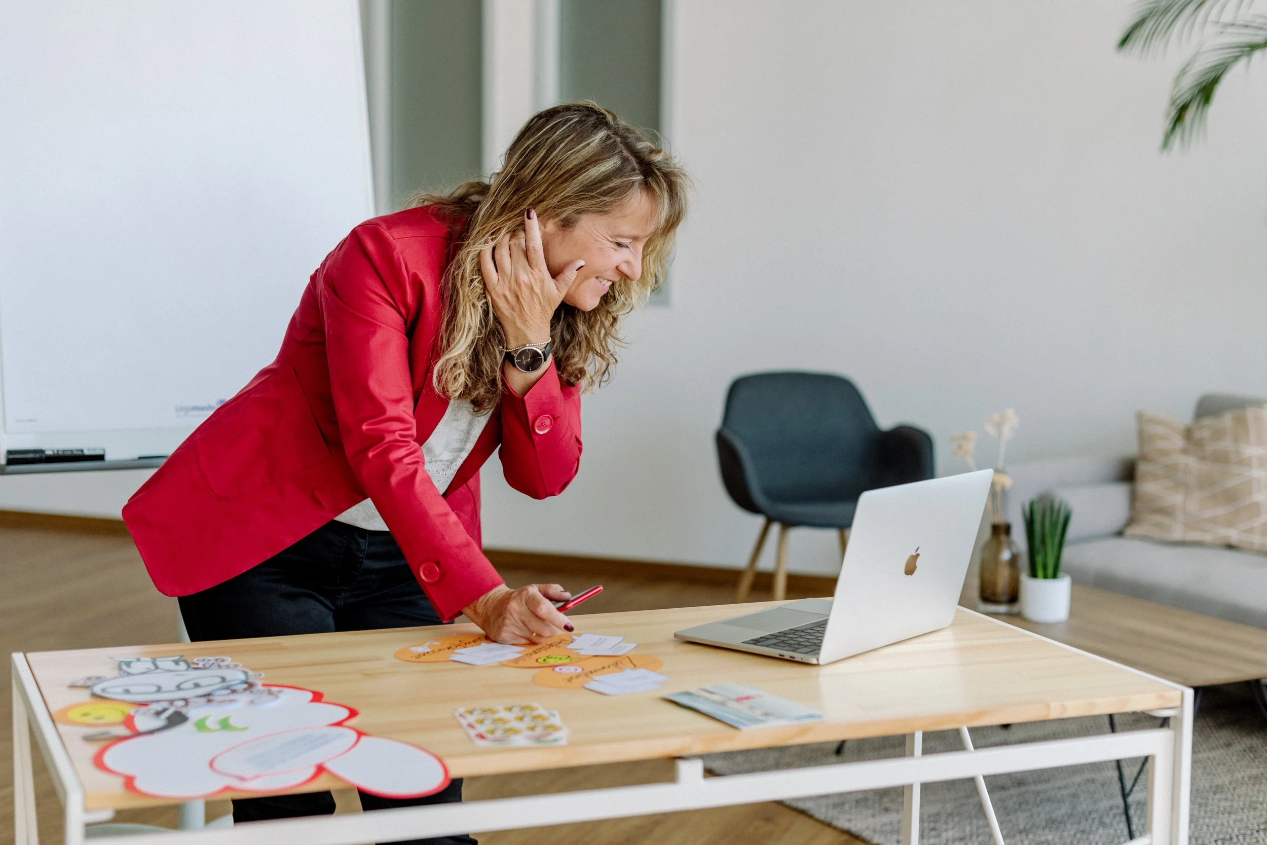 Eine lachende Frau in rotem Blazer arbeitet an einem Tisch mit einem Laptop und bunten Bastelmaterialien in einem modernen Büroraum.