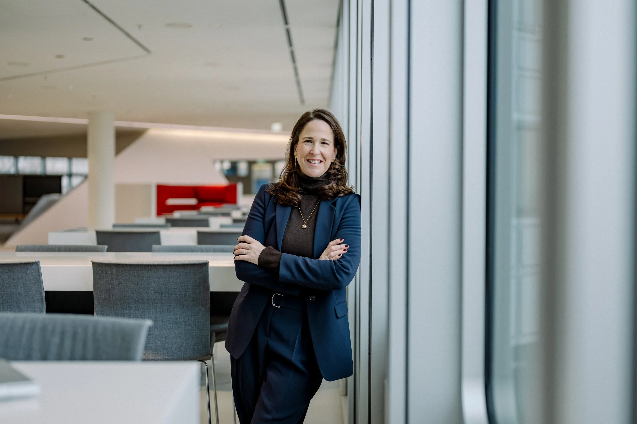 Eine lächelnde Frau mit dunklem Haar, die eine dunkelblaue Jacke trägt, steht in einem modernen, lichtdurchfluteten Büro mit großen Fenstern und ansprechend gestalteter Einrichtung.