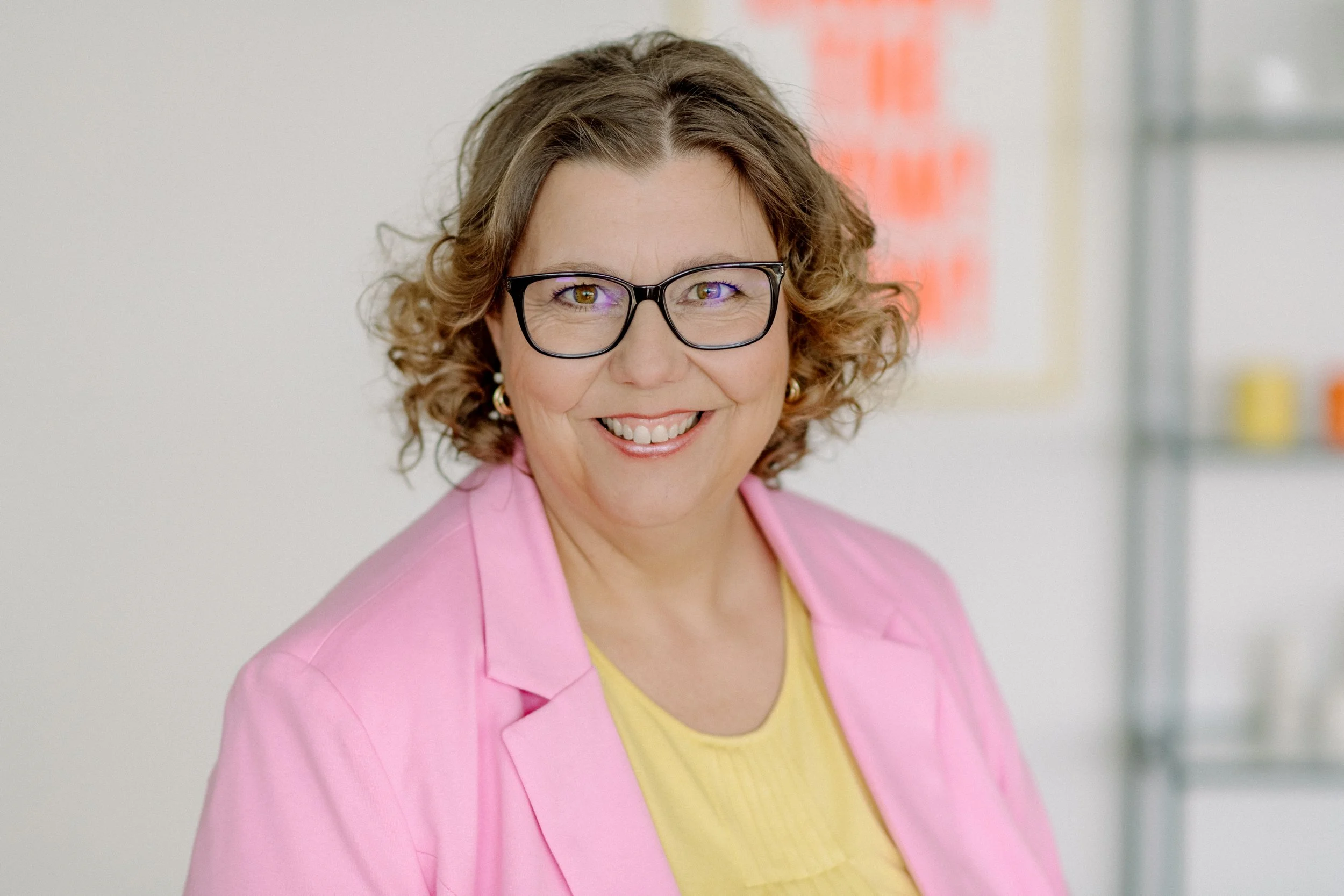 Eine lächelnde Frau mit Brille trägt eine pinke Jacke und eine gelbe Bluse, im Hintergrund eine weiße Wand mit rot markiertem Poster.