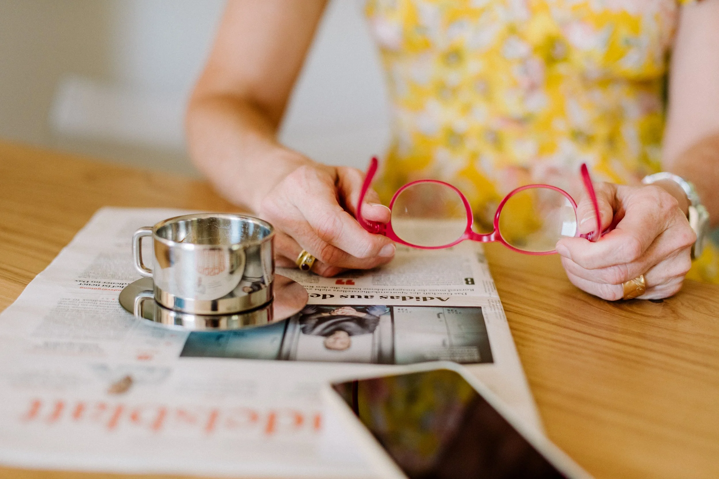 Person hält eine rote Brille in der Hand, auf dem Tisch liegt ein Zeitungsausschnitt, im Vordergrund ein metallischer Becher.