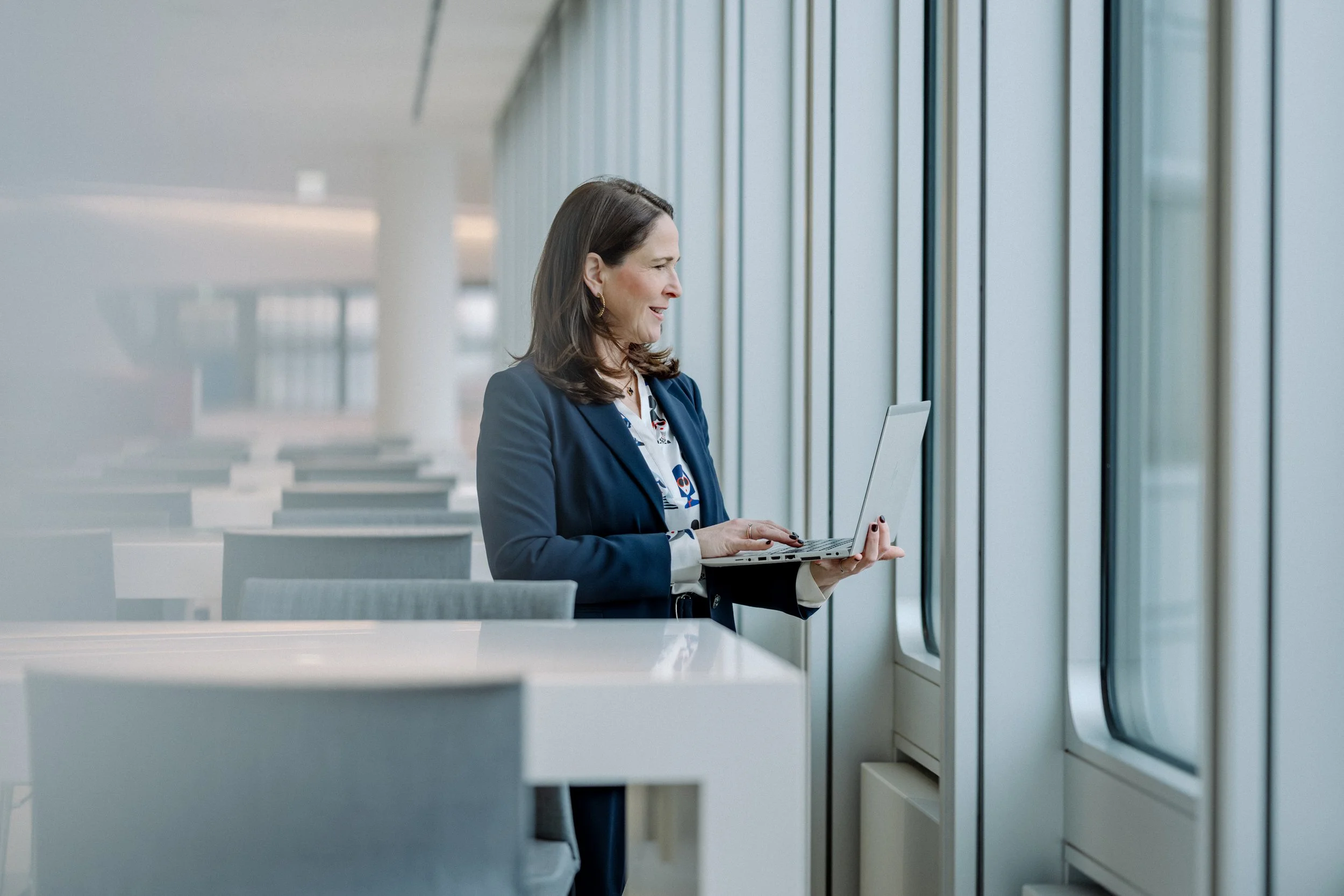 Eine Frau mit dunklem Haar, die in einem modernen, hellen Gebäude an einem Laptop arbeitet, neben einem großen Fenster.