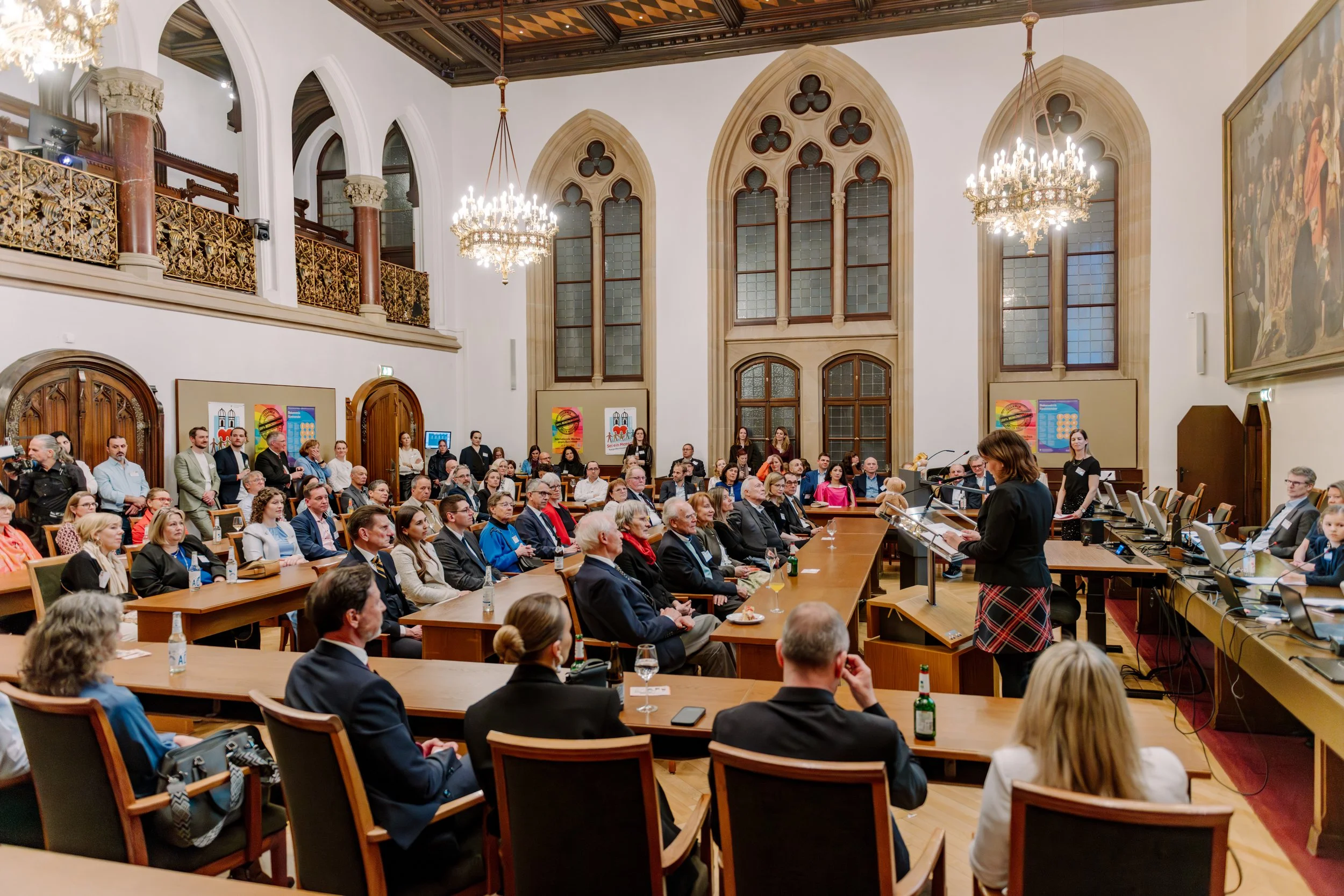 Ein großer Saal mit hohen Fenstern im gotischen Stil, in dem ein Treffen oder eine Konferenz mit vielen Menschen stattfindet. Es gibt Kronleuchter, Bänke und Tische, an denen die Teilnehmer sitzen. Eine Frau spricht vorne am Rednerpult.