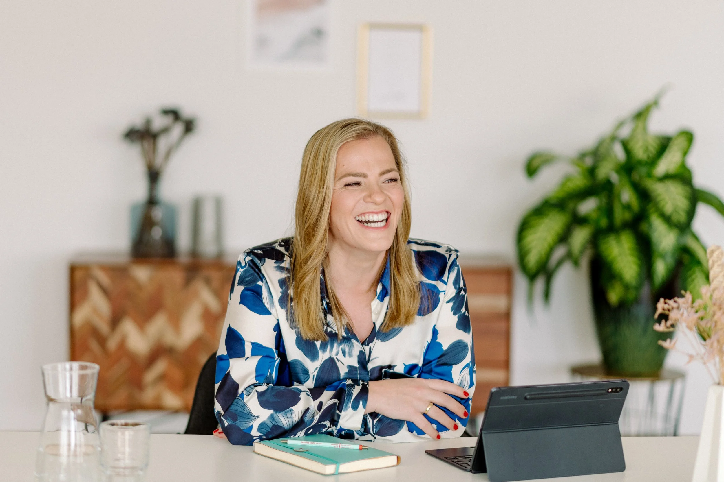 Eine lachende Frau mit blondem Haar, die an einem Tisch mit einem Laptop und Notizbüchern sitzt, umgeben von Pflanzen und Dekorationen in einem modernen Raum.
