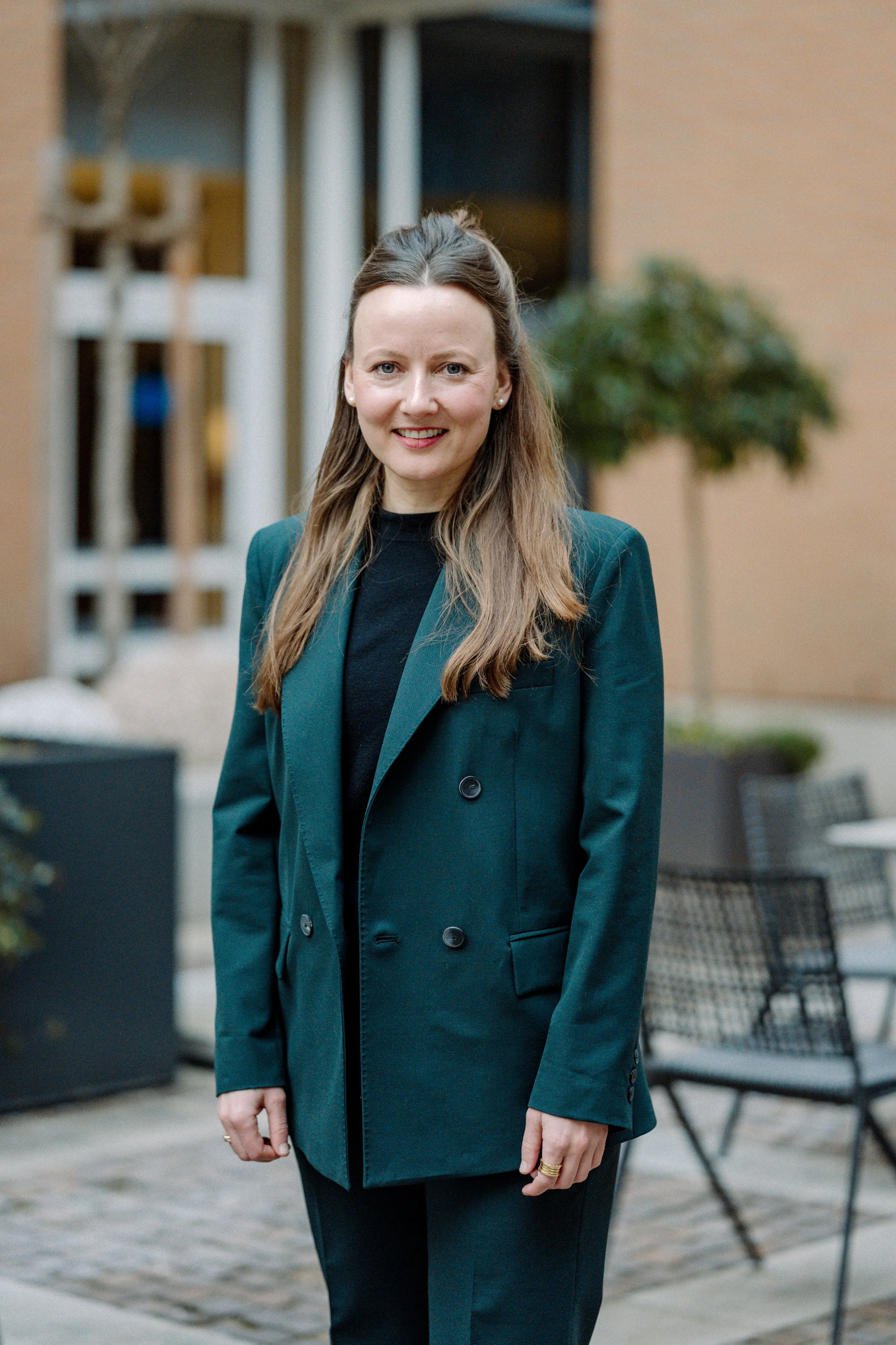 Porträt einer Frau in einem schwarzen Blazer, die draußen vor einem modernen Gebäude steht.